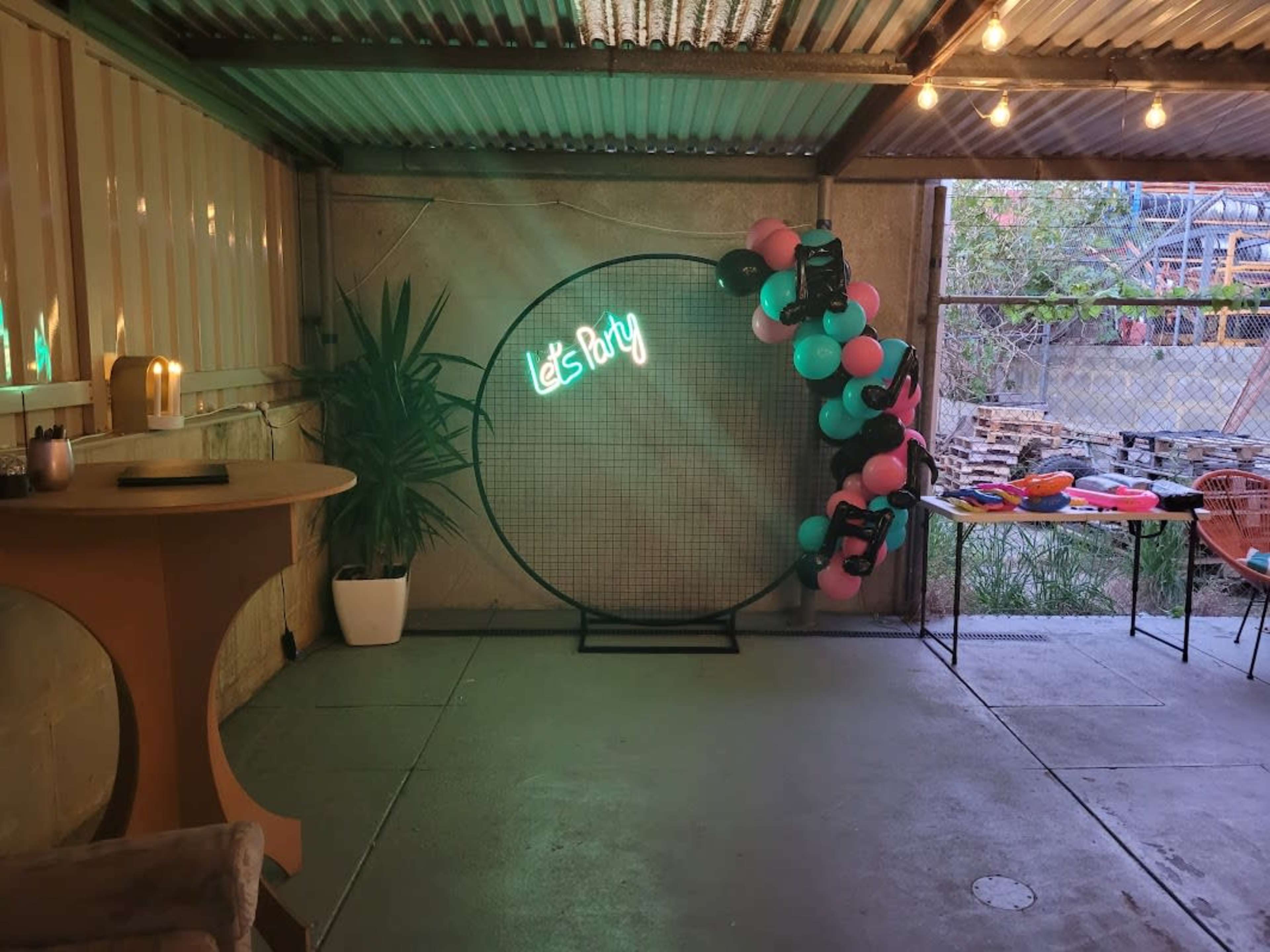 A partially decorated indoor space featuring a round netted backdrop with a neon sign that says "Let's Party," surrounded by colorful balloons and a small table in the foreground.