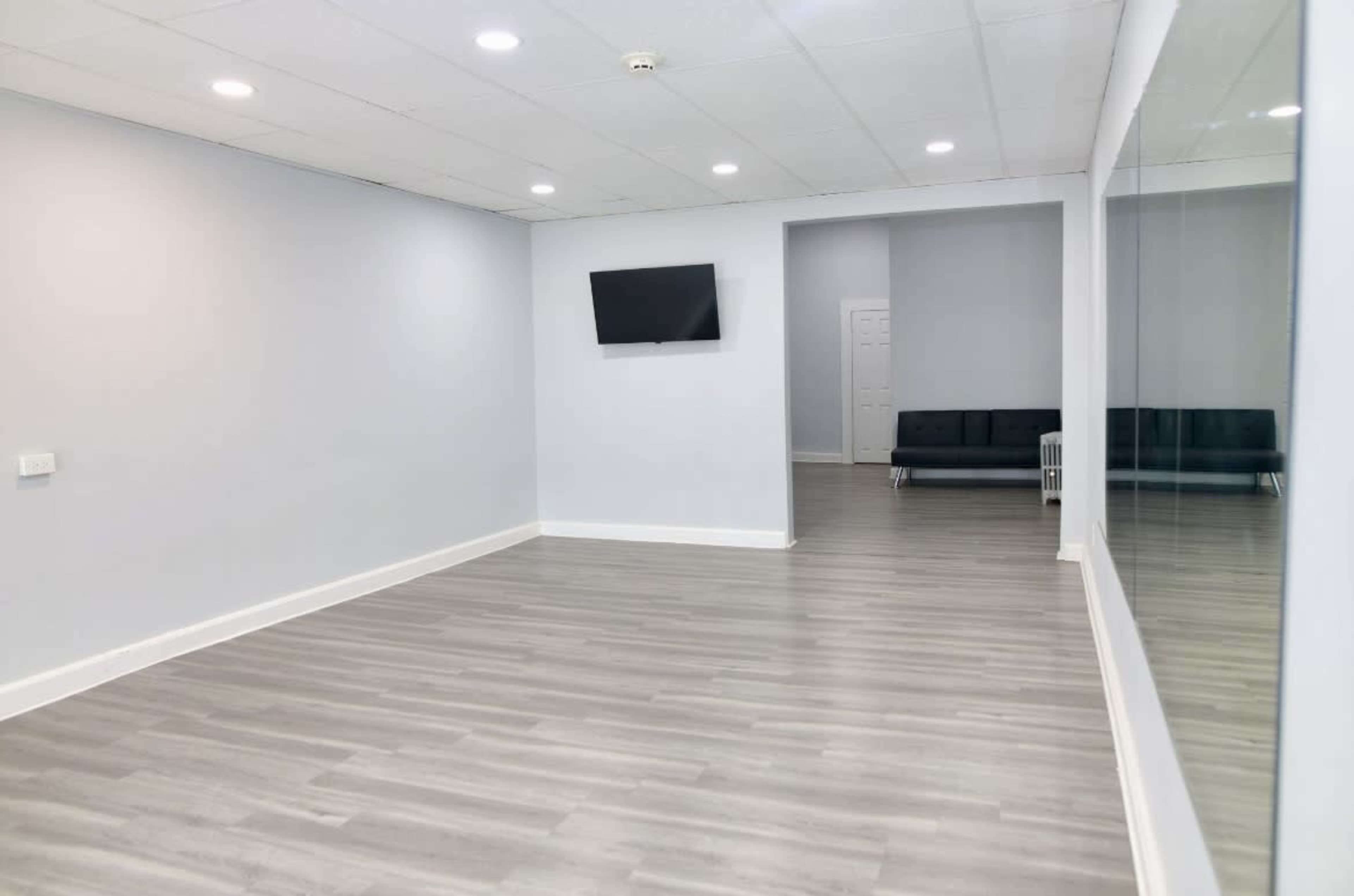 A spacious, light-colored room features a wall-mounted television, a mirrored wall, and a minimalist seating area in the background.