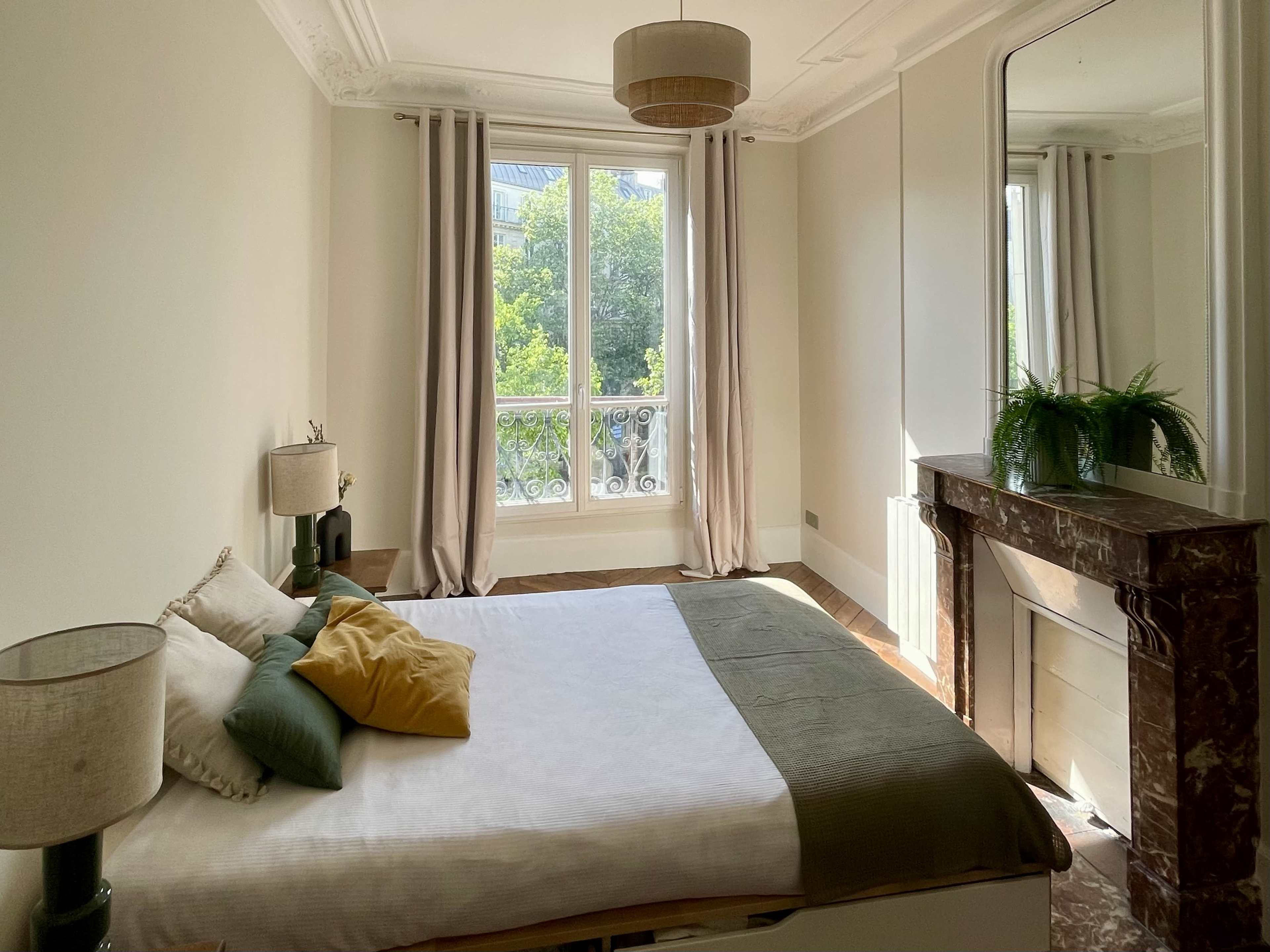 A bright bedroom features a bed with decorative pillows, a marble fireplace, two lamps, and large windows draped with light curtains.