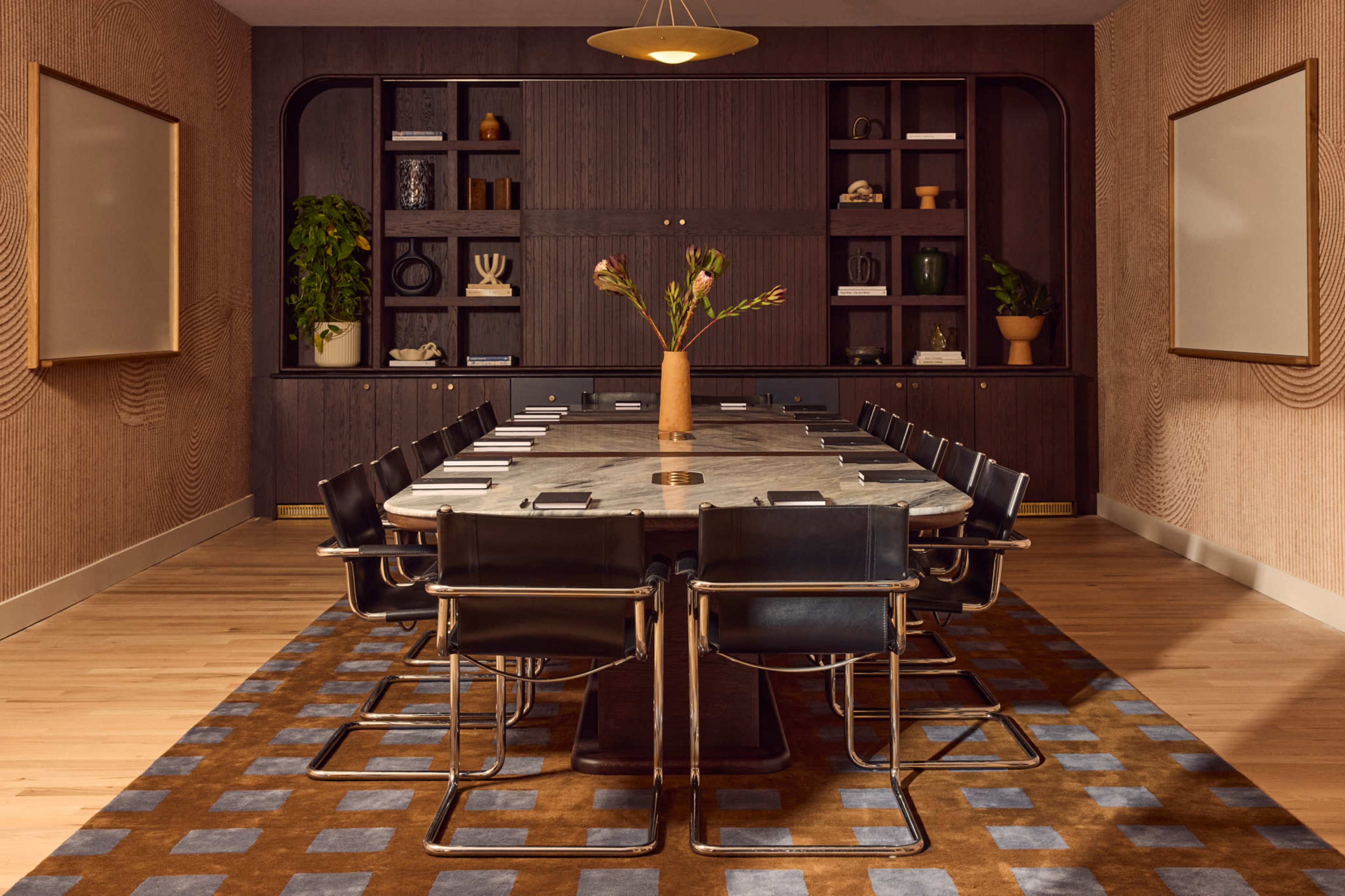 A modern conference room features a large marble table surrounded by black chairs, with a floral centerpiece and a backdrop of shelves filled with decorative items and plants.