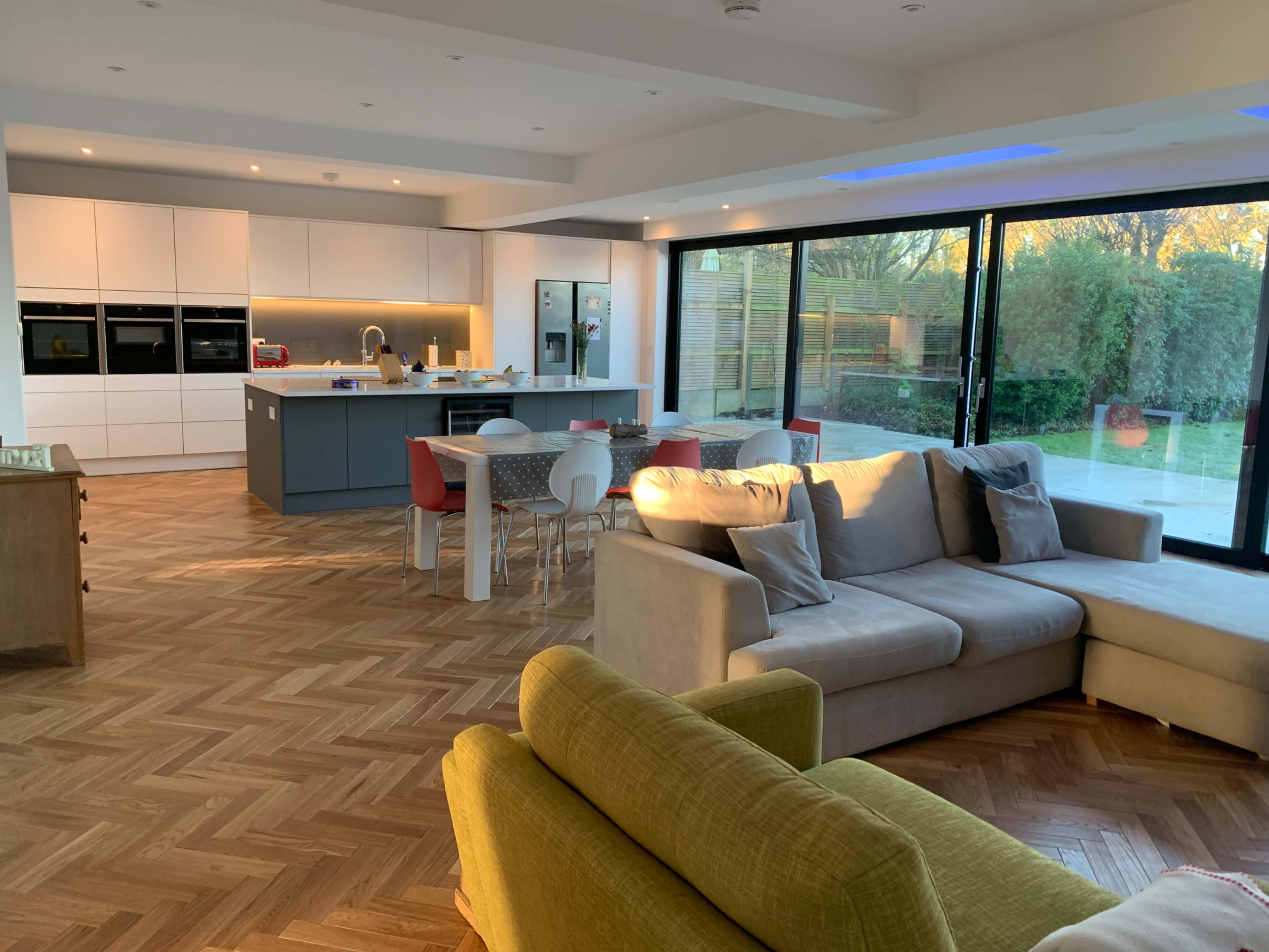 A modern kitchen and living area feature a large sectional sofa, a dining table with colorful chairs, and floor-to-ceiling windows overlooking a garden.