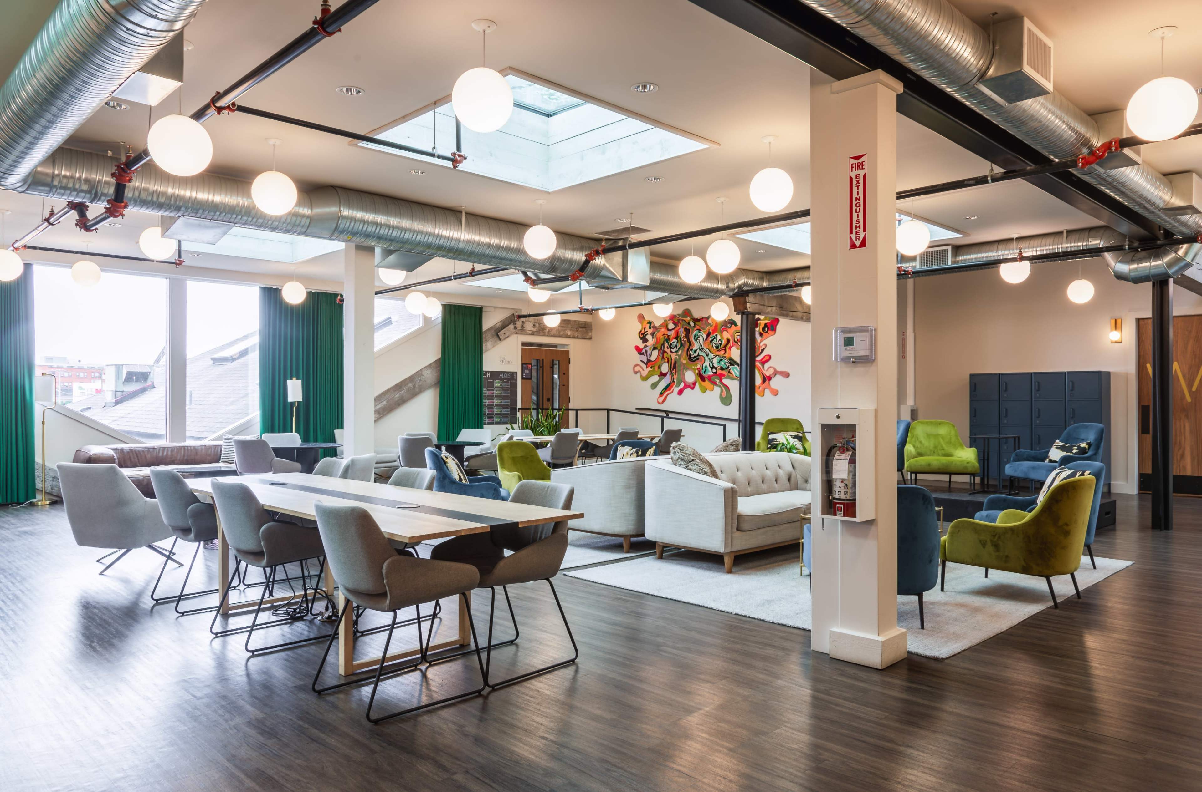 The image shows a modern, open lounge area featuring a mix of seating options, a large table, and colorful wall art, with ample natural light from skylights.