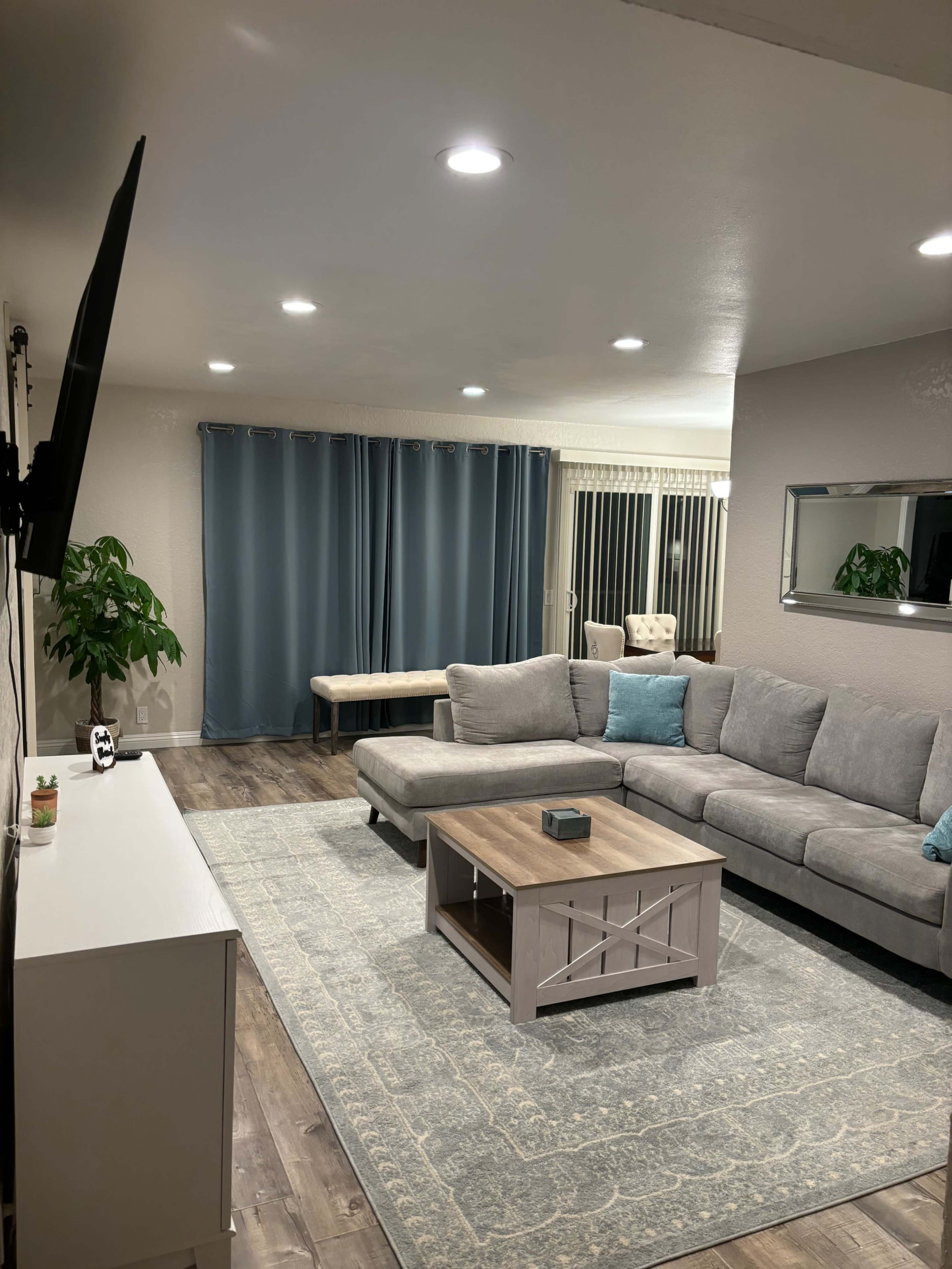 A modern living room features a large gray sectional sofa, a wooden coffee table, and blue curtains, with a television mounted on the wall.