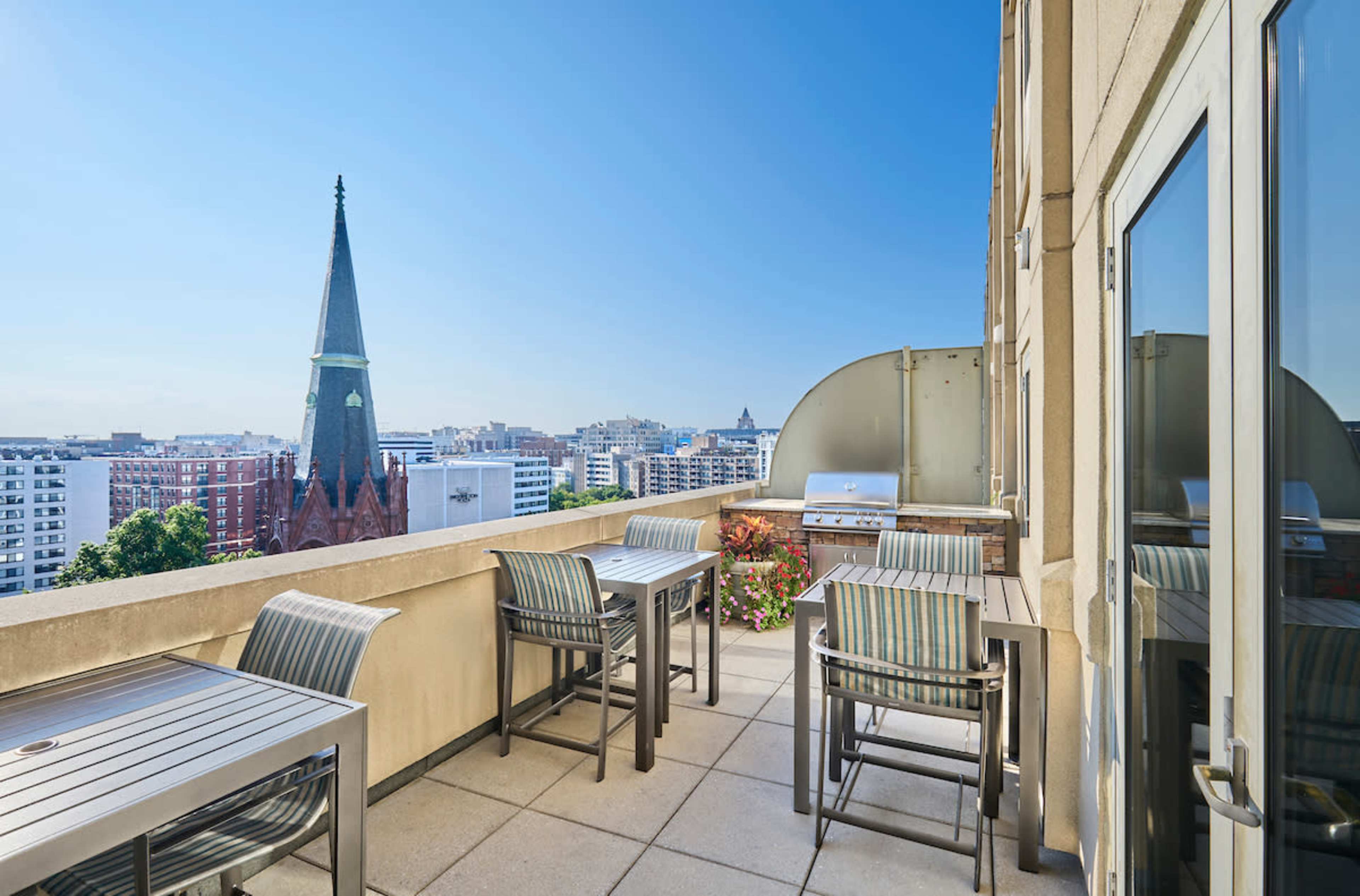 A balcony features metallic tables and chairs, overlooking a city skyline with a prominent spire in the background.