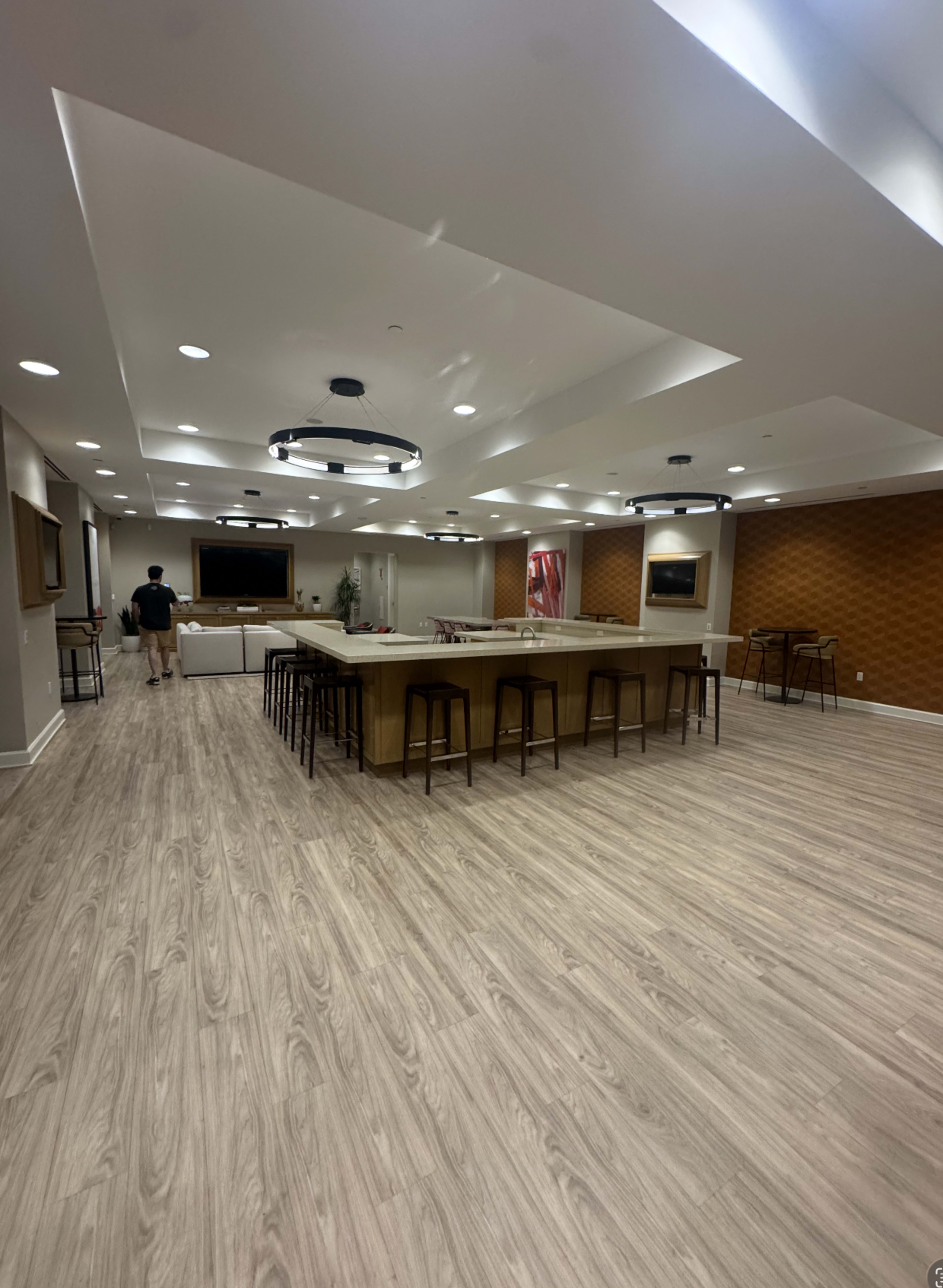 The image shows a spacious, modern room with wooden flooring, a long kitchen island, and a seating area, accompanied by a person walking in the background.