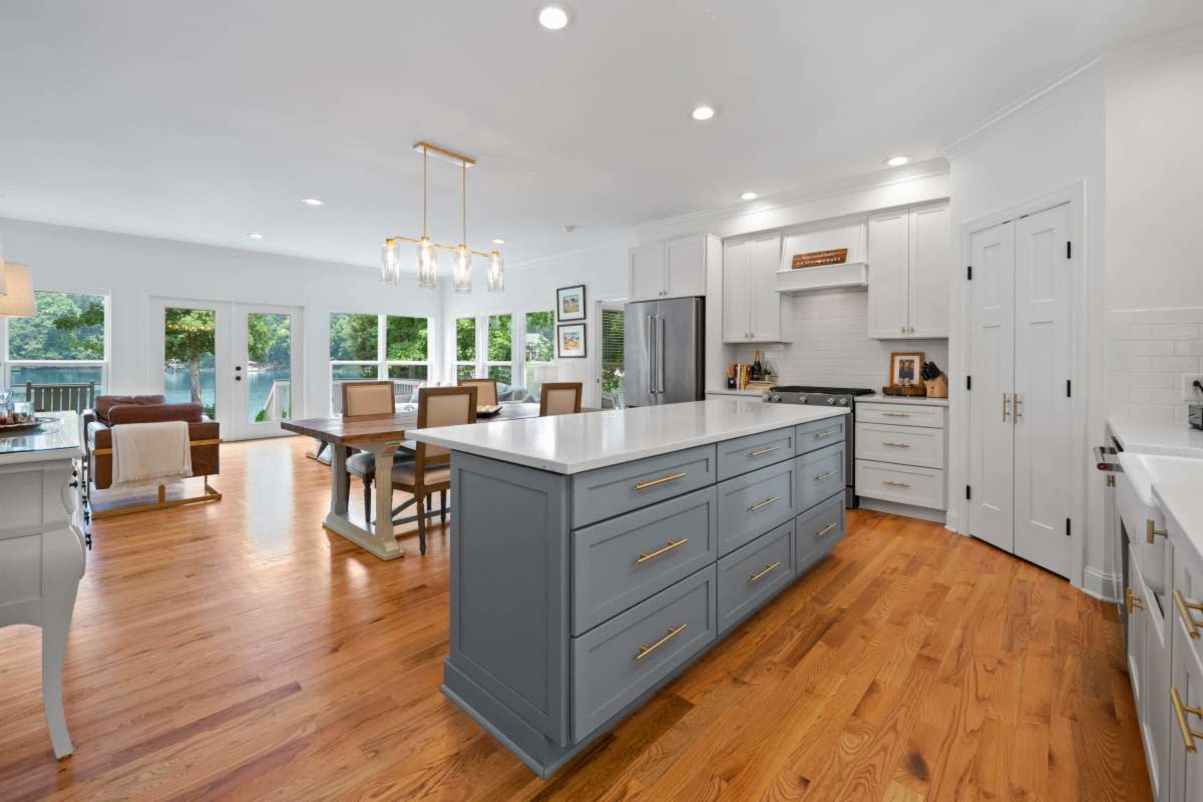 The image shows a modern kitchen featuring a large central island with a gray finish, surrounded by wooden cabinetry and dining furniture, with large windows letting in natural light.