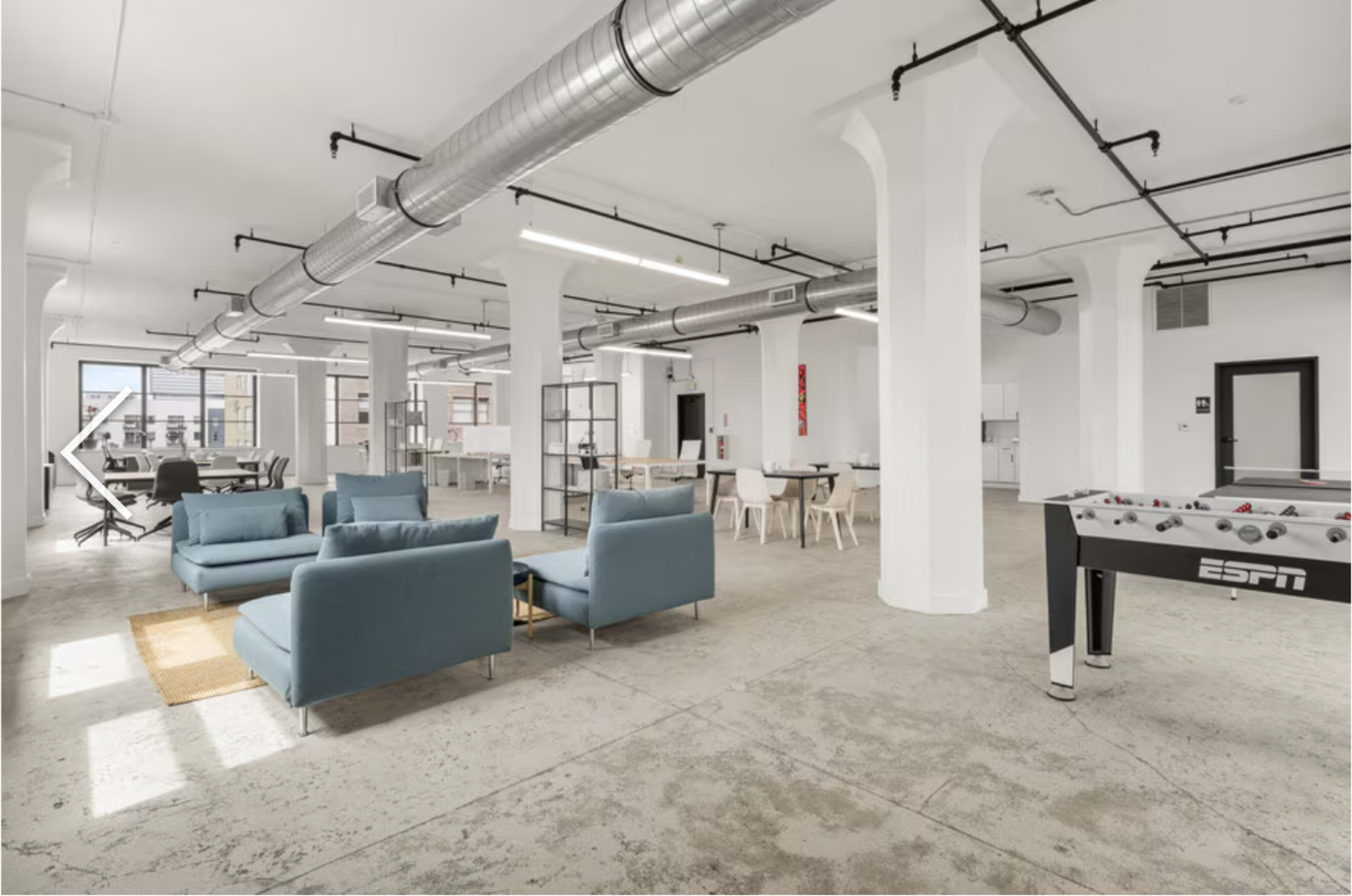 The image shows a spacious, modern office with light-colored walls, large windows, and a mix of work and relaxation areas, including blue sofas and a foosball table.