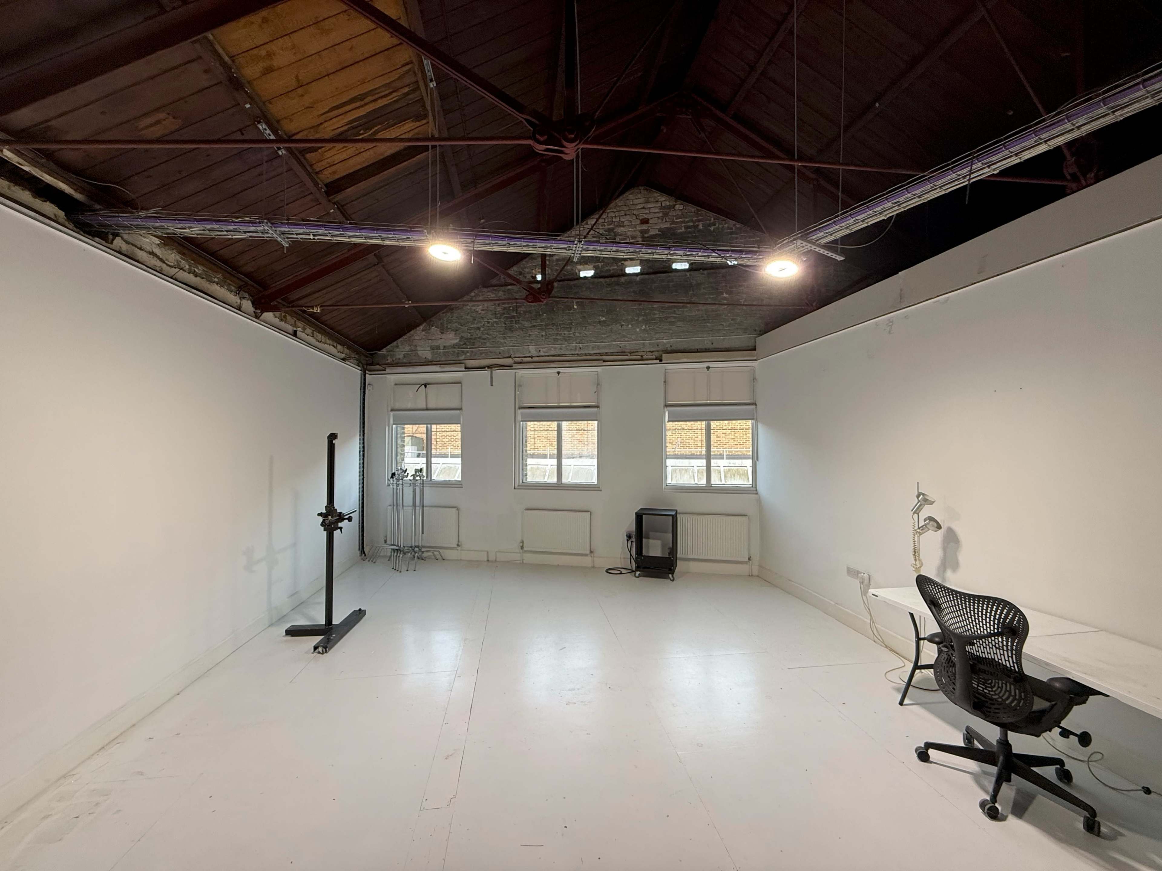 The image shows a spacious, empty room with a high ceiling, white walls, and minimal furnishings, including a chair and a black stand.