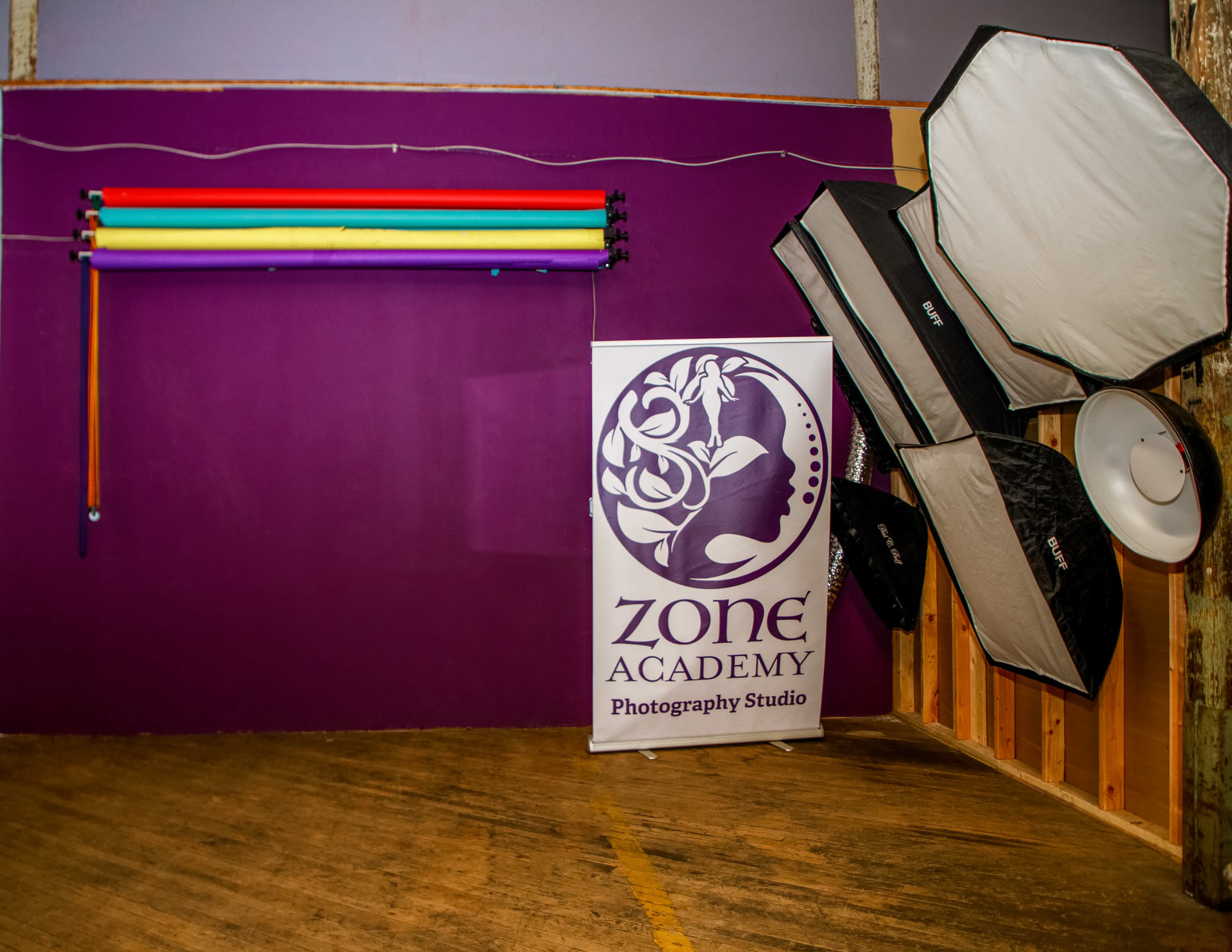 The image shows the interior of a photography studio featuring a purple wall with a banner for "Zone Academy Photography Studio" and various photography lighting equipment on the right.