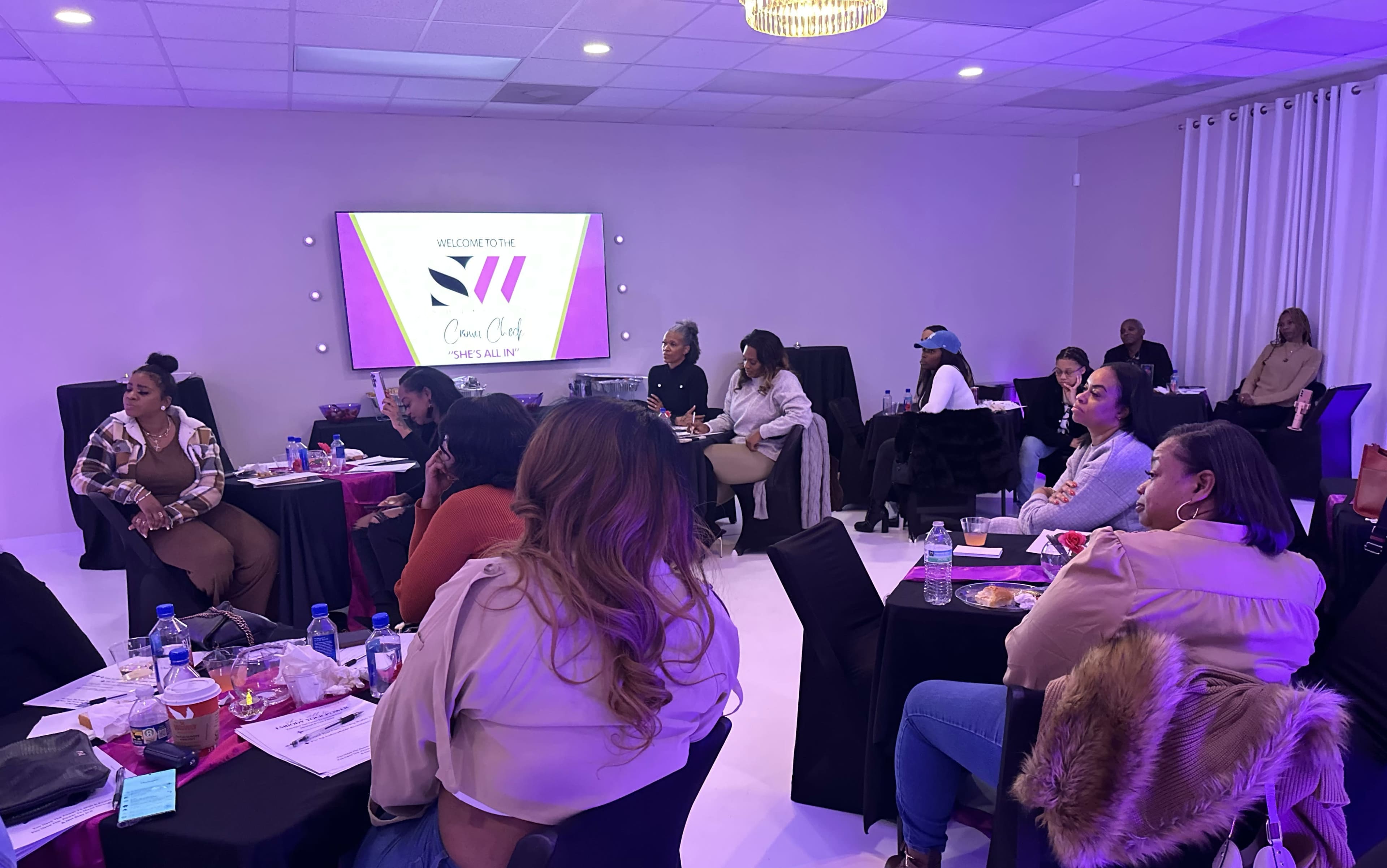 A group of people sits at tables in a purple-lit room during a meeting or event, facing a screen with a welcome message.