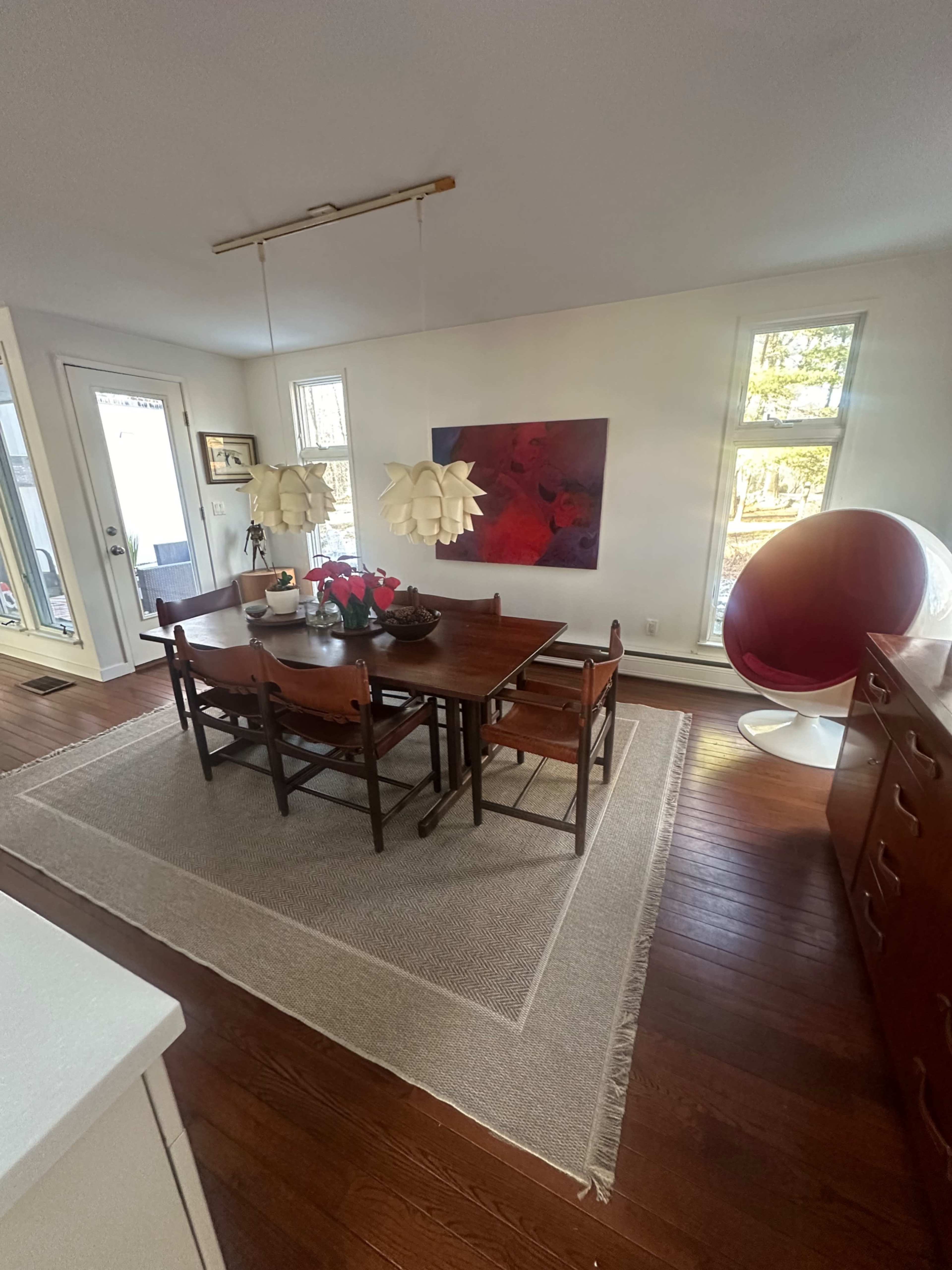 A dining area features a wooden table surrounded by chairs, a modern light fixture above, and a large painting on the wall, with a window and a round chair in the corner.