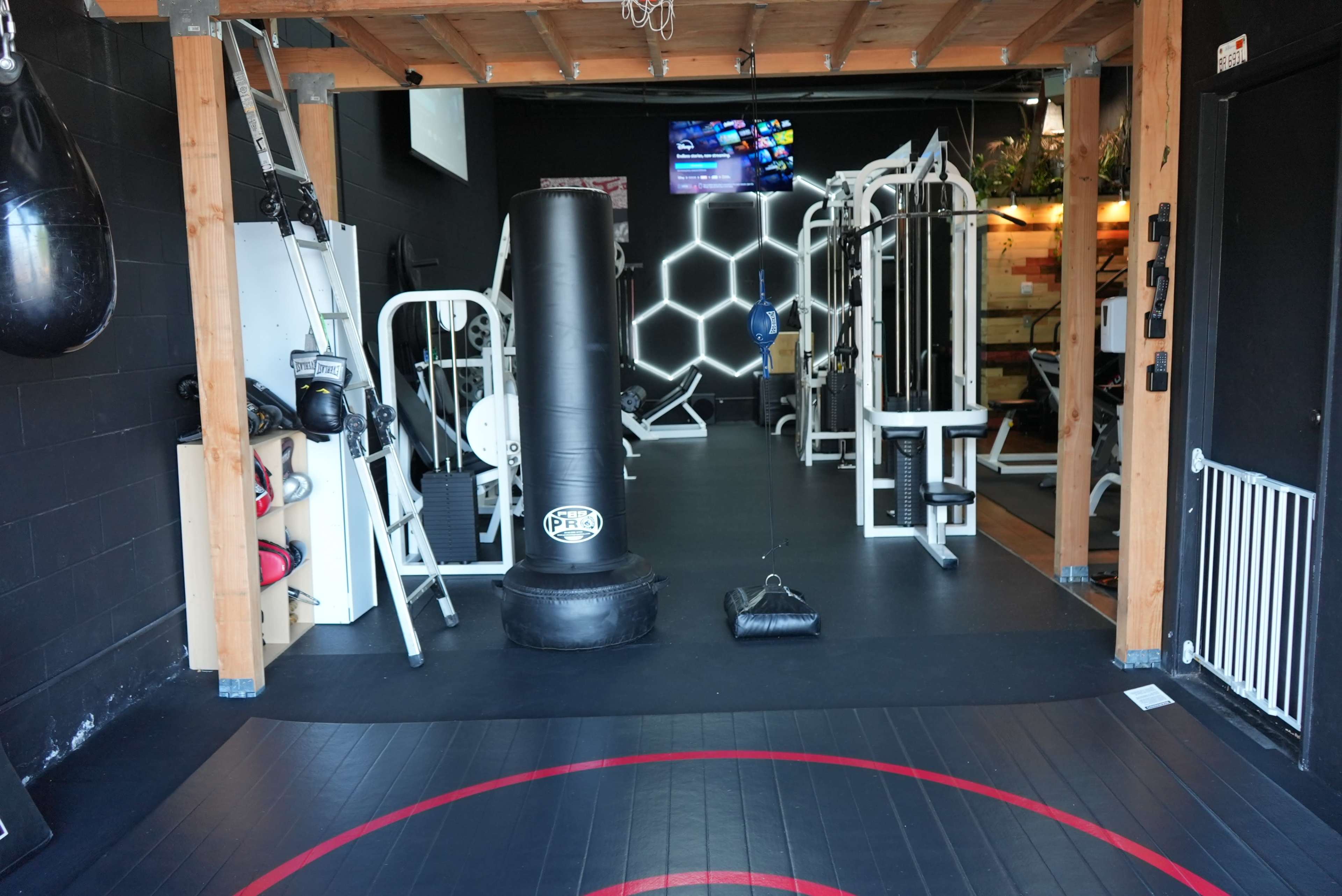 A gym interior featuring various workout equipment, including a heavy bag, a punching bag, and weight machines, under a wooden ceiling and illuminated by a television screen on the wall.