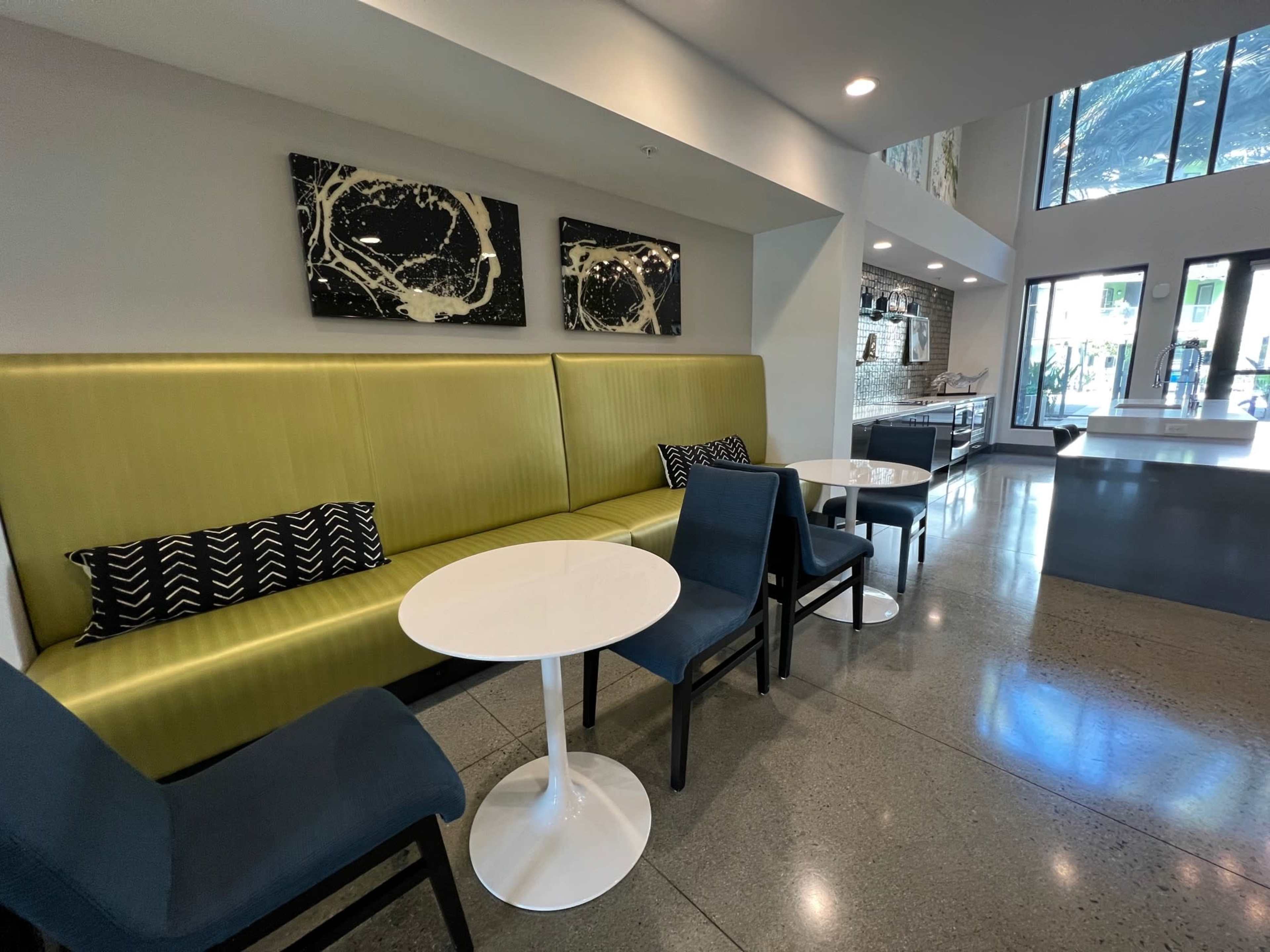 A modern seating area with a green upholstered bench, white tables, and dark blue chairs under large windows.