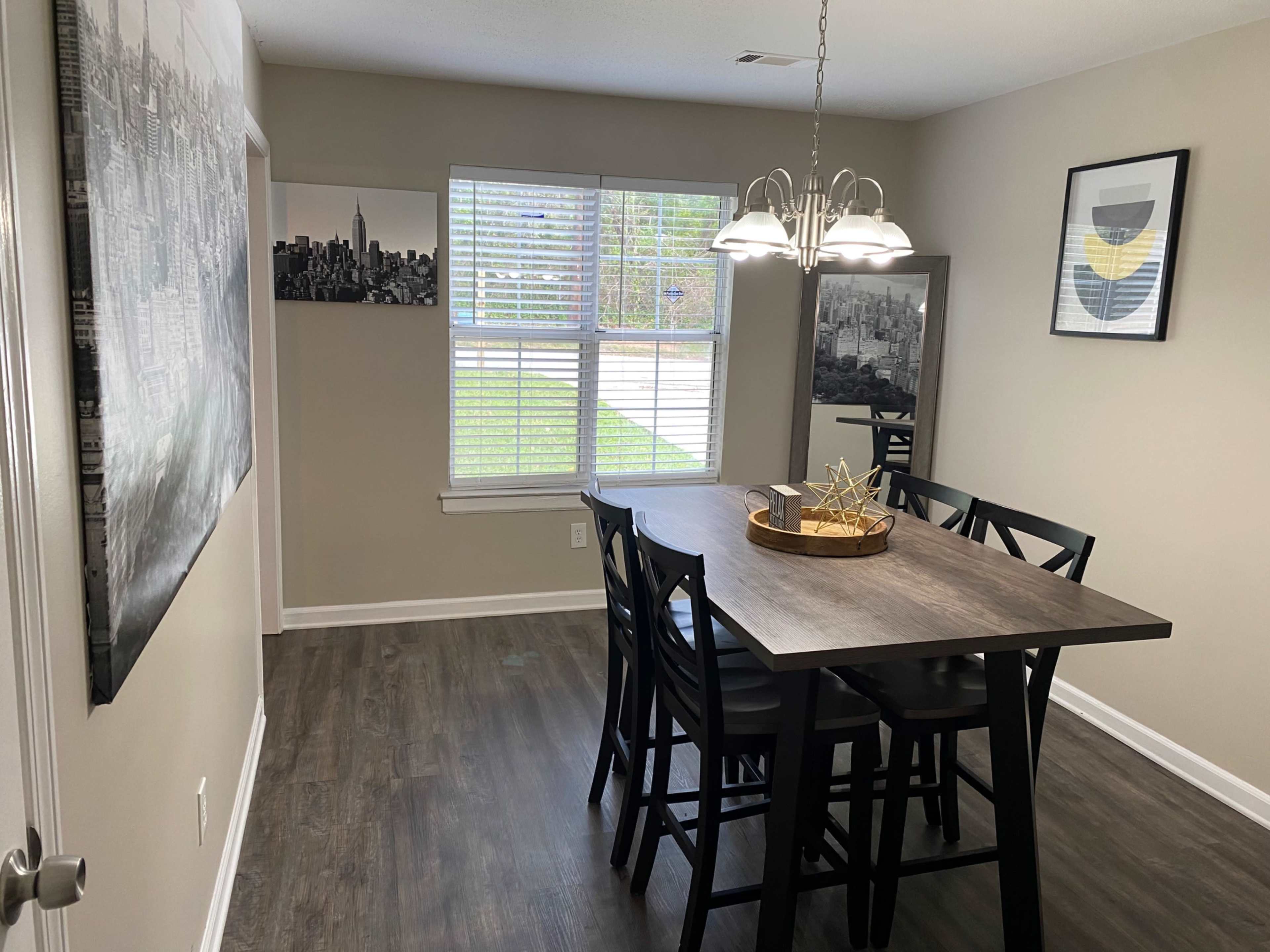 A dining area features a wooden table surrounded by black chairs, with light fixtures above and large windows letting in natural light.