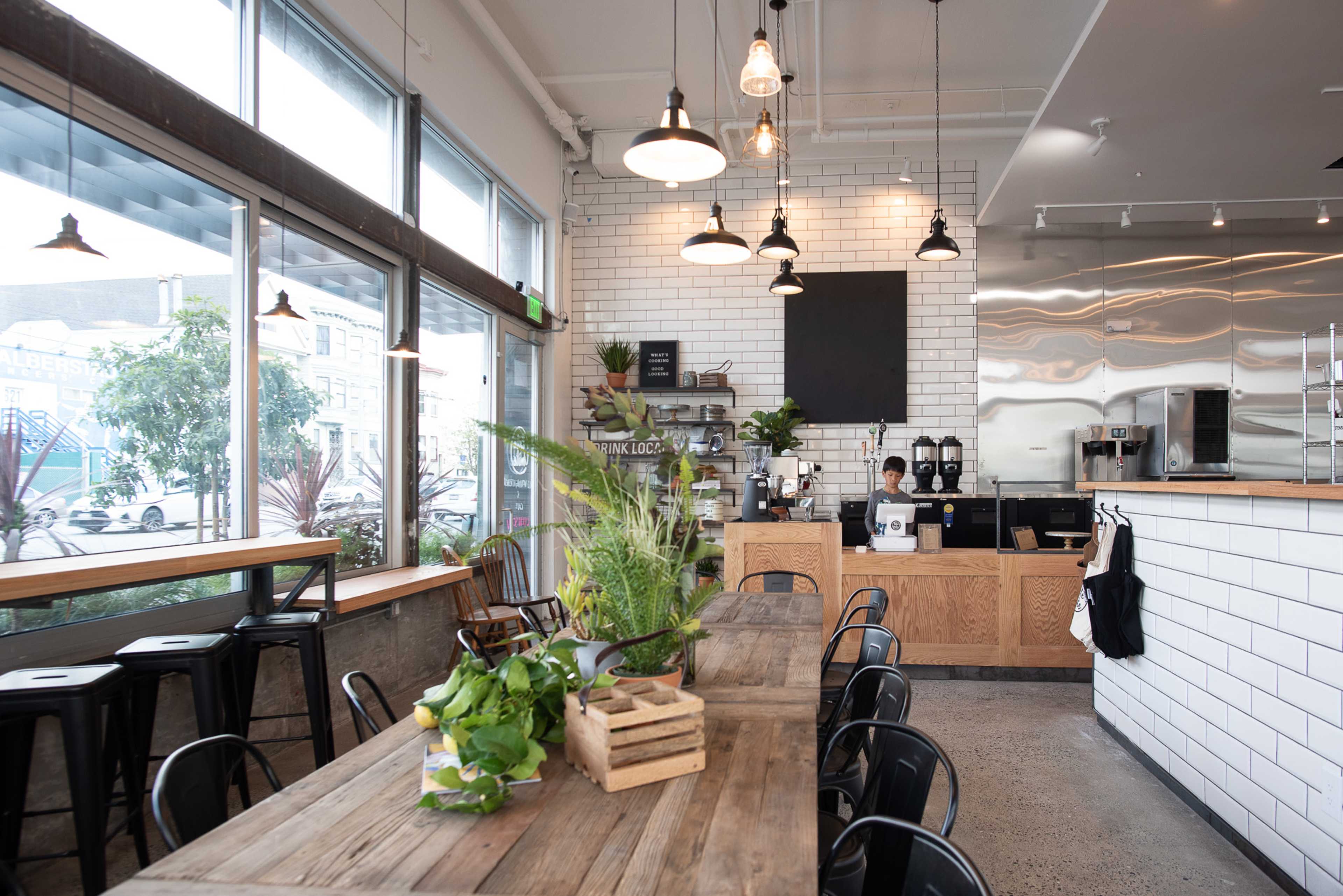 A modern café features a wooden communal table, industrial-style seating, and a sleek counter with coffee equipment against a backdrop of large windows.