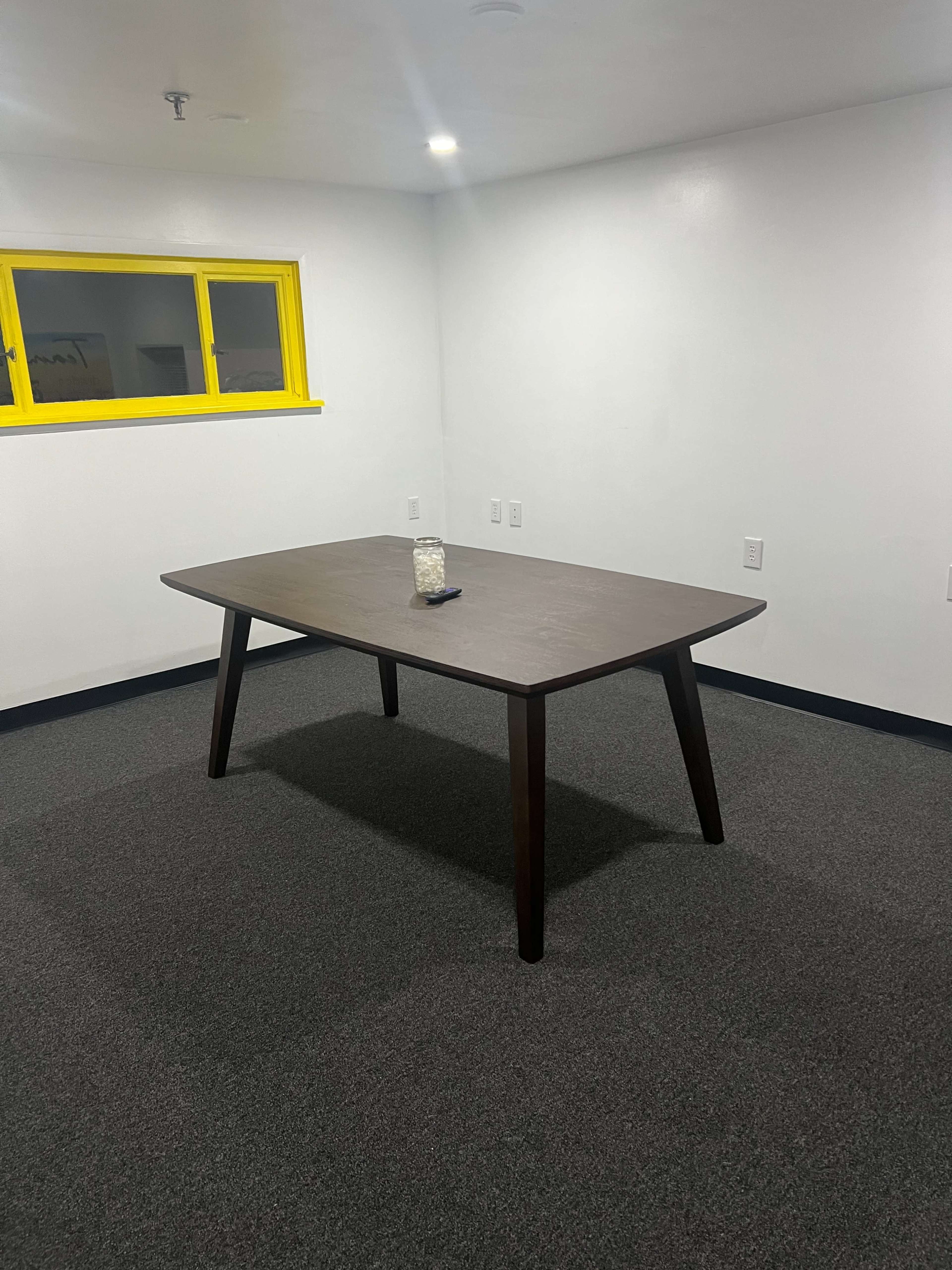 A wooden table with angled legs is positioned in the center of a sparsely furnished room with gray carpeting and a brightly colored yellow window.