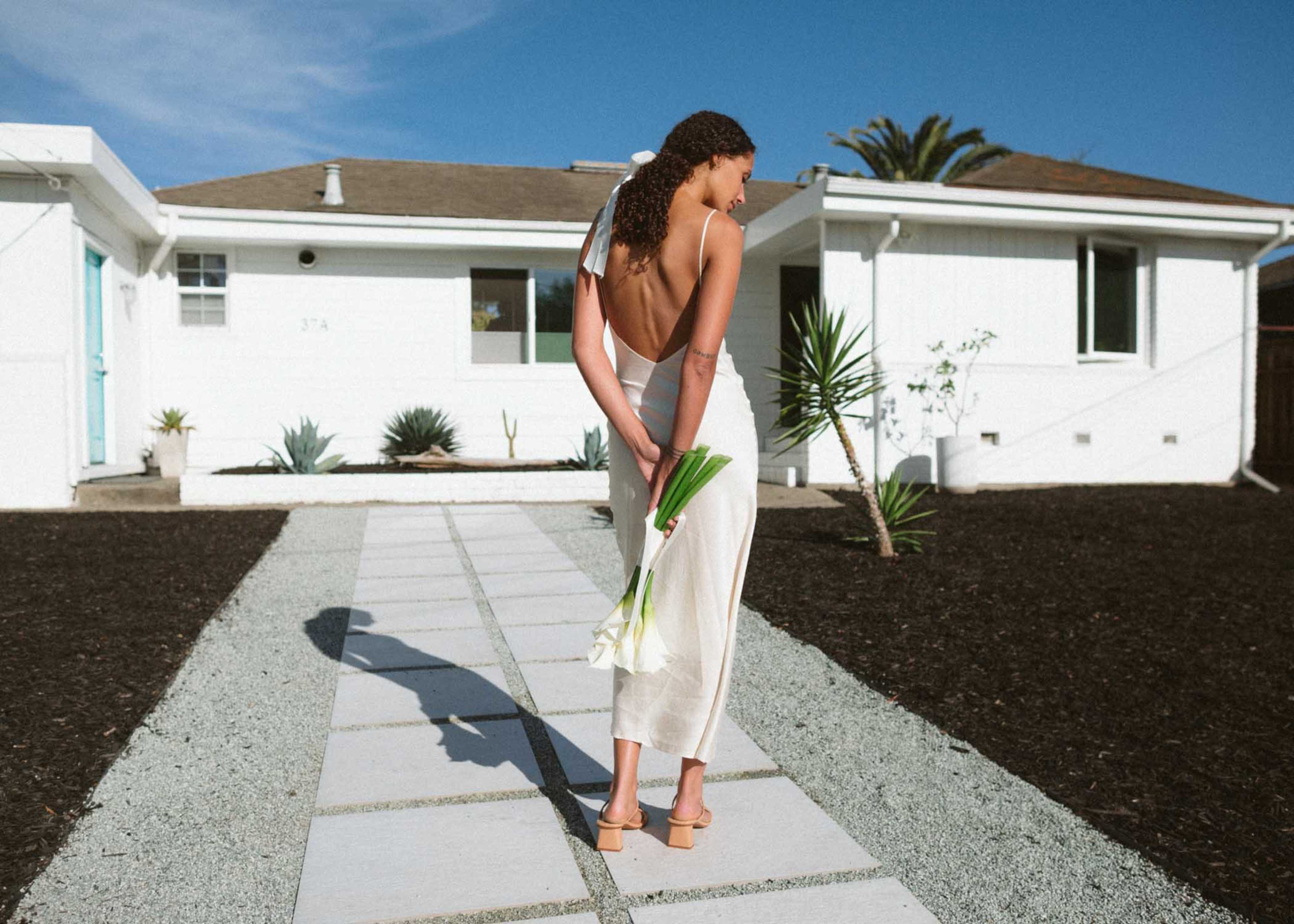 A woman stands barefoot on a stone pathway, holding white flowers behind her back in front of a white house with a blue door and palm trees.