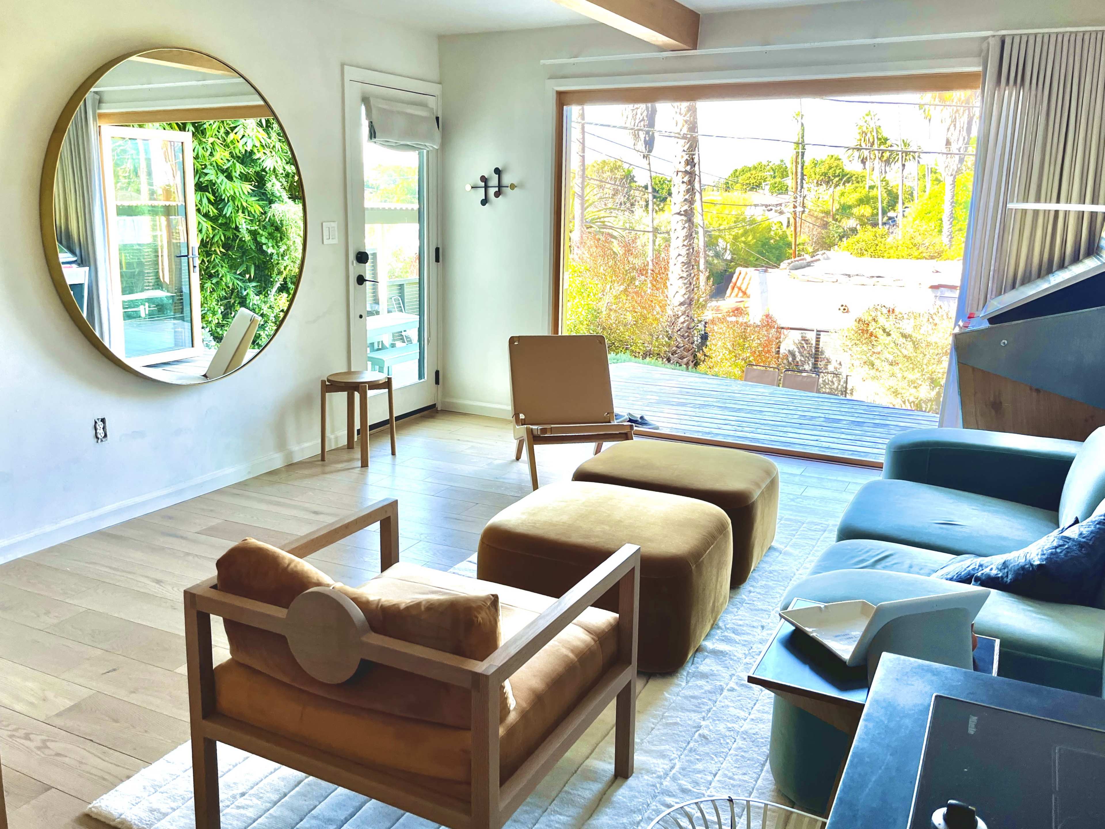 A bright living room features a round mirror, a blue sofa, wooden chairs, and a view of greenery through large windows.