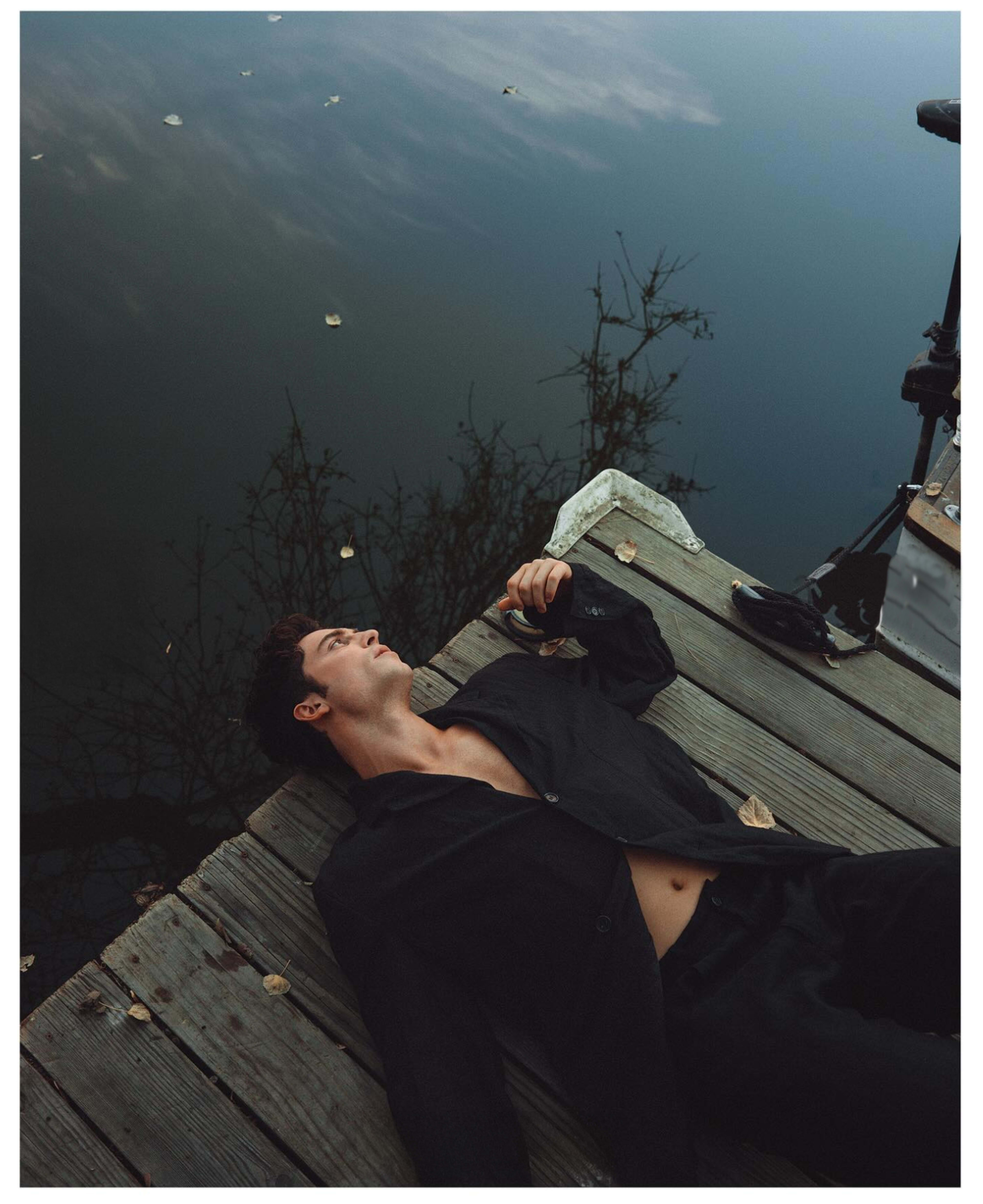 A man lies on a wooden dock near calm water, gazing upward with leaves scattered around him.