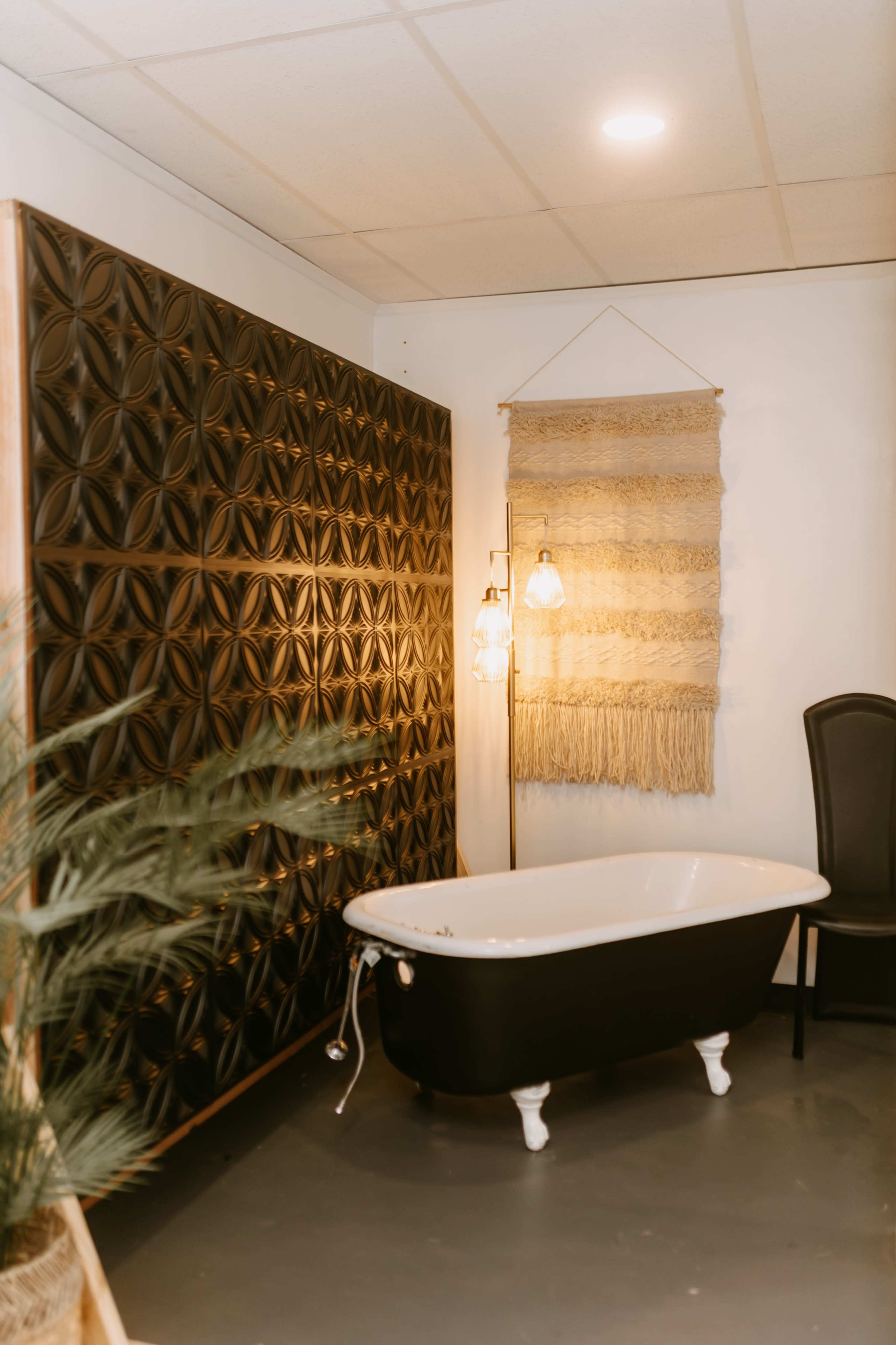 The image shows a stylish bathroom featuring a black and white freestanding bathtub, a textured black wall, a woven wall hanging, a floor lamp, and a chair.