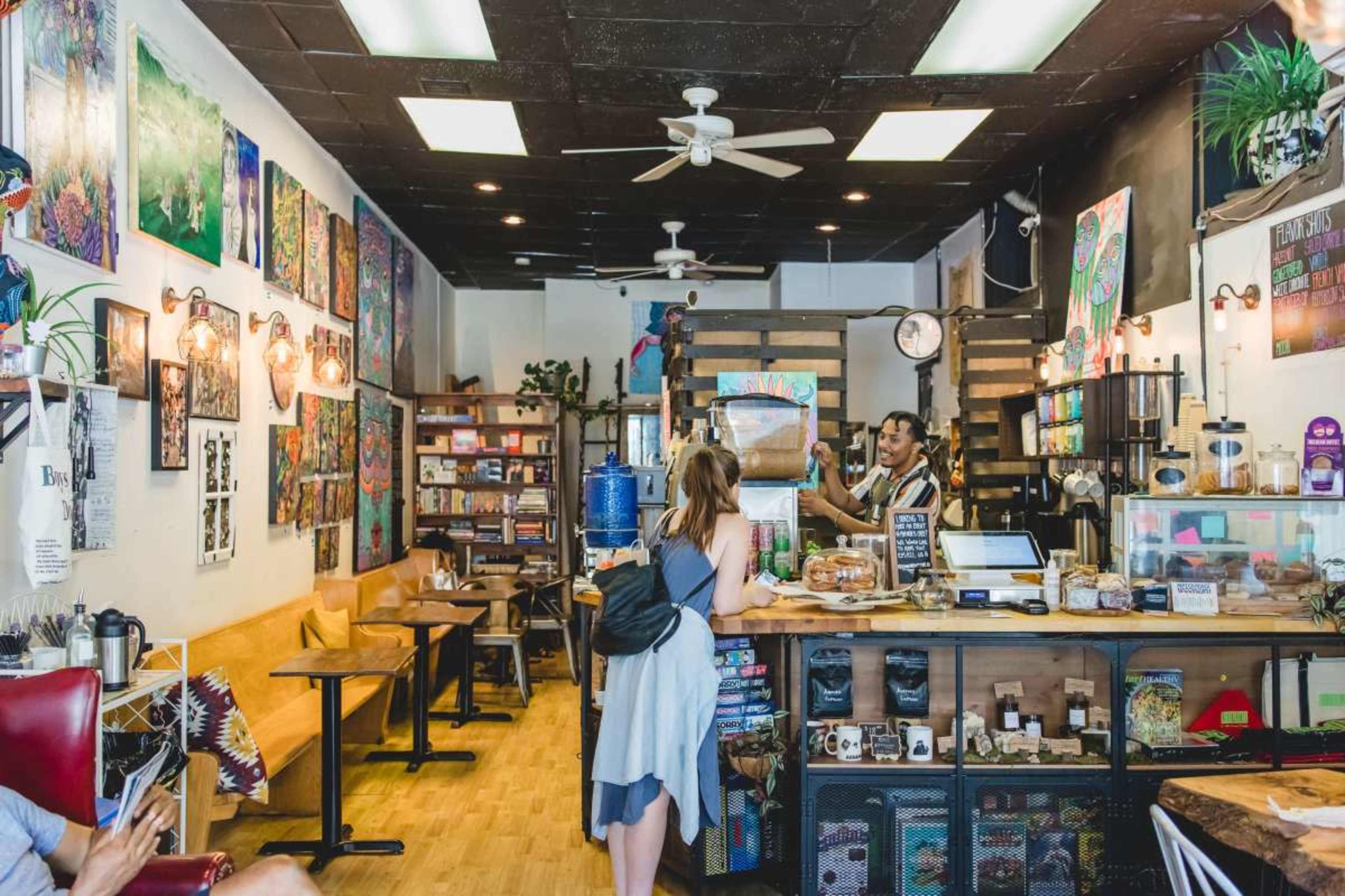 A woman with long hair stands at a counter in a colorful café filled with artwork and wooden seating.