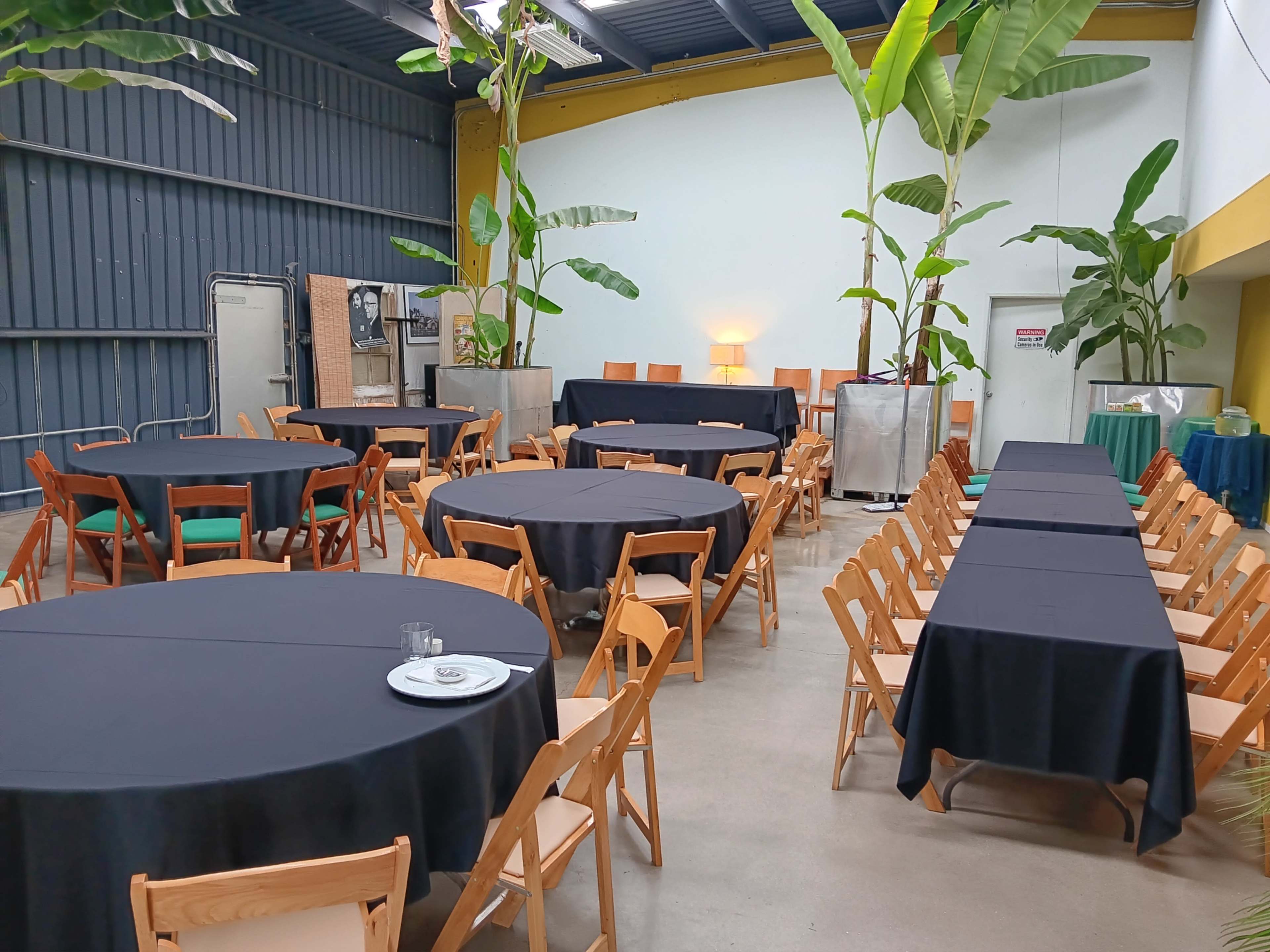 A spacious indoor venue features several round and rectangular tables covered with black tablecloths, surrounded by wooden chairs and decorative plants.