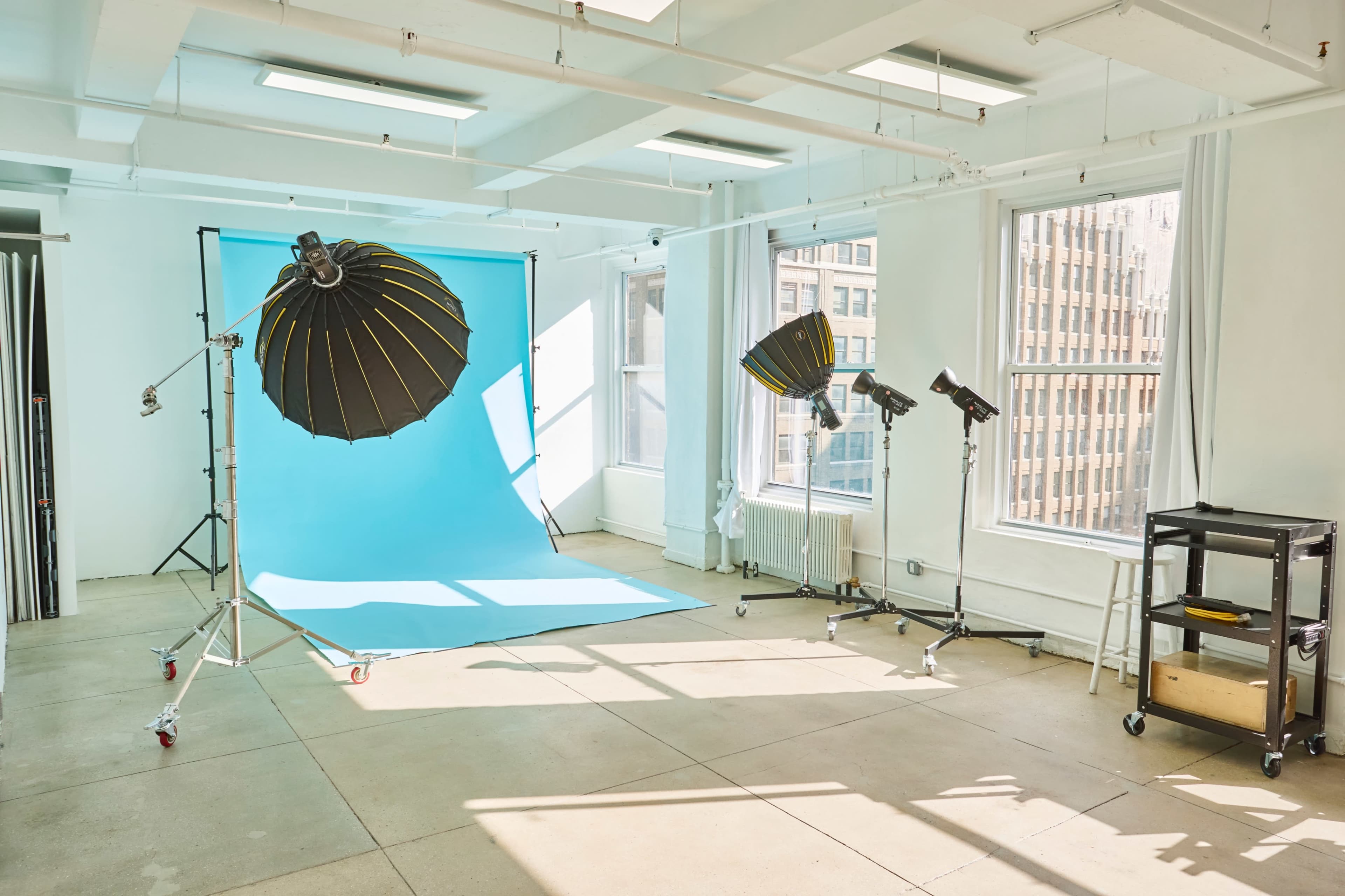 A photography studio features a bright blue backdrop, three softbox lights, and large windows allowing natural light to flood the space.
