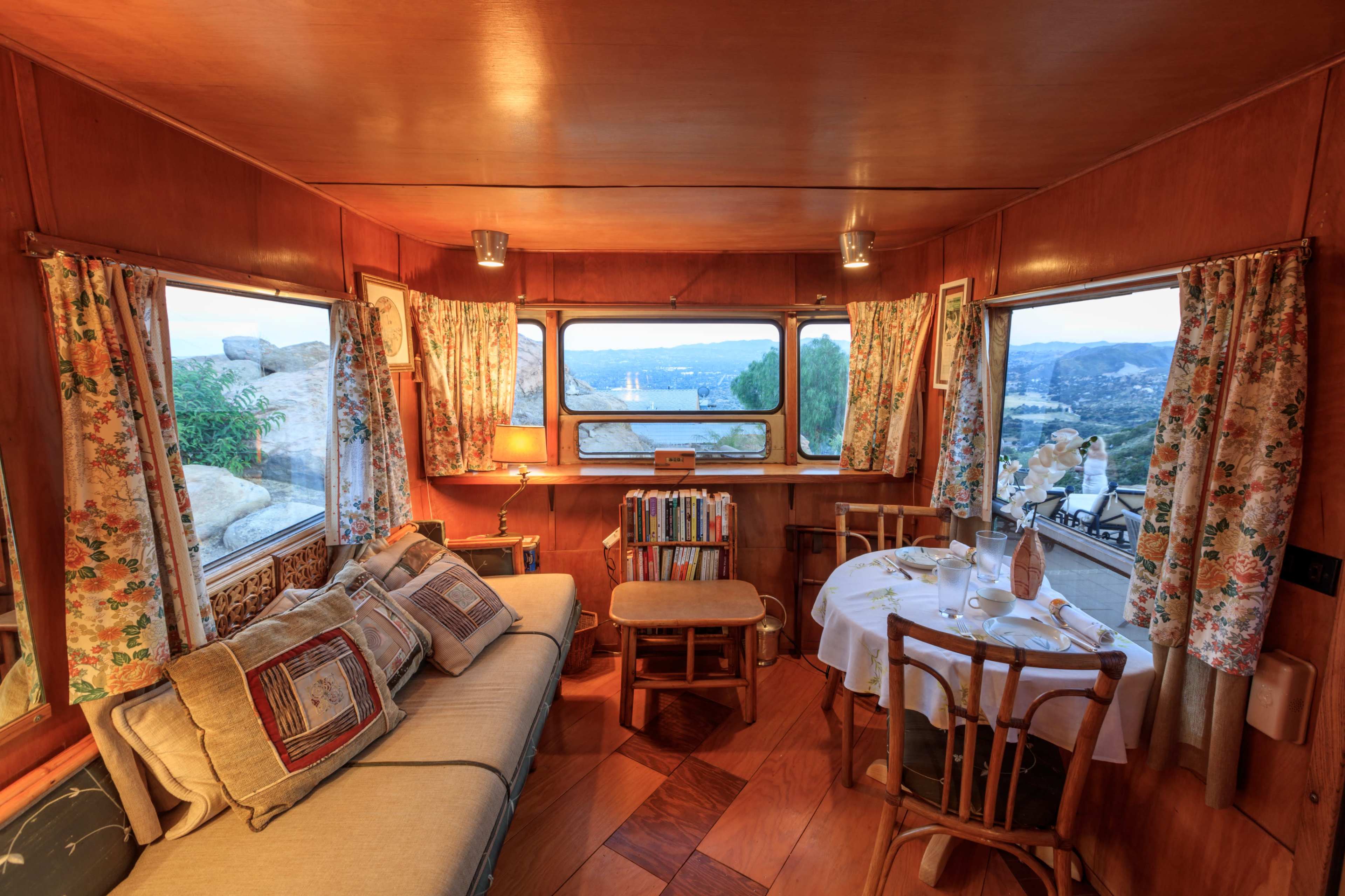 A cozy interior of a rustic cabin features a dining area, a sofa, and large windows showcasing a scenic view.