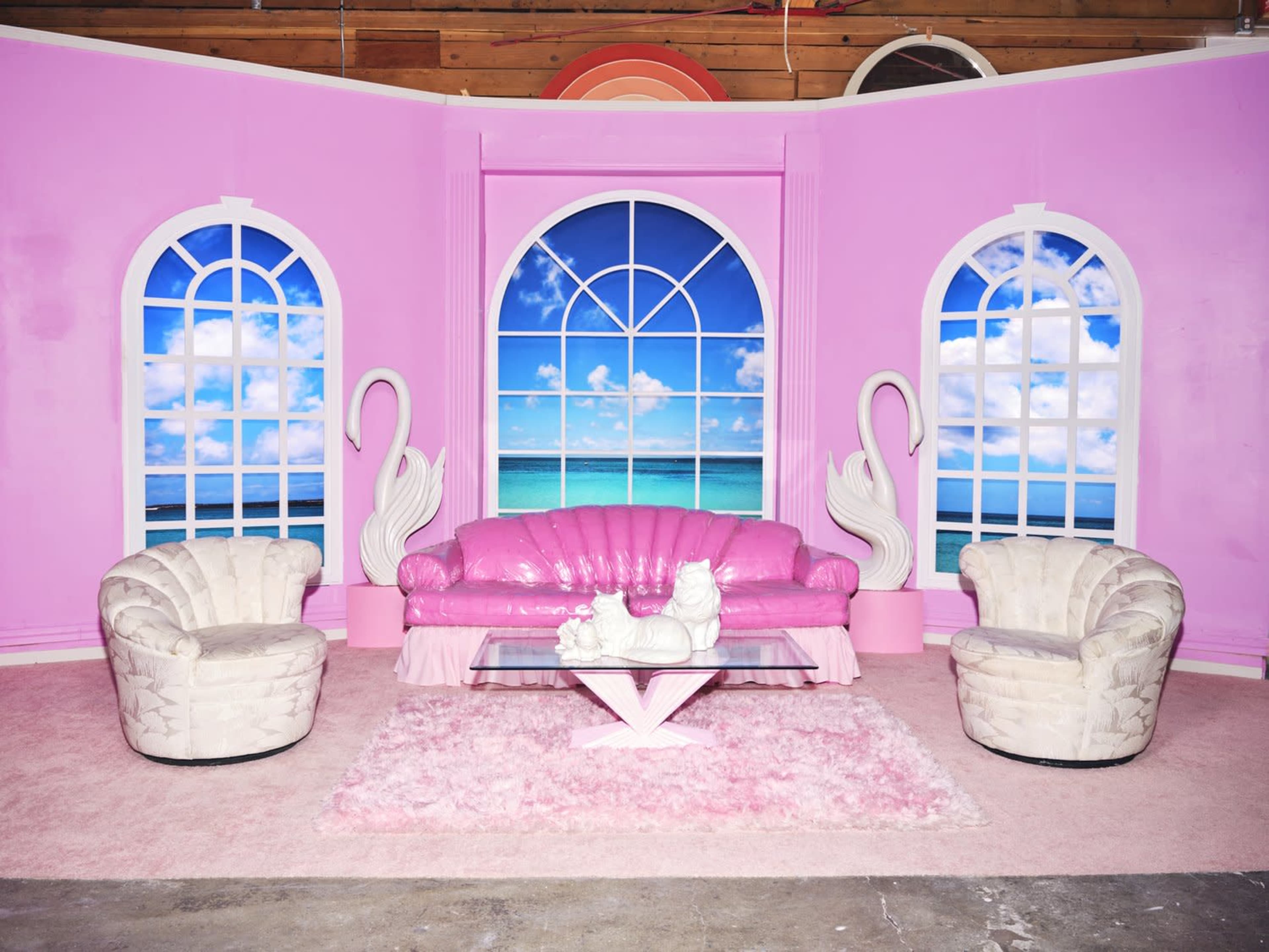 The scene features a pink room with large arched windows showing a beach view, furnished with a pink sofa, two white chairs, and a glass coffee table on a fluffy area rug.