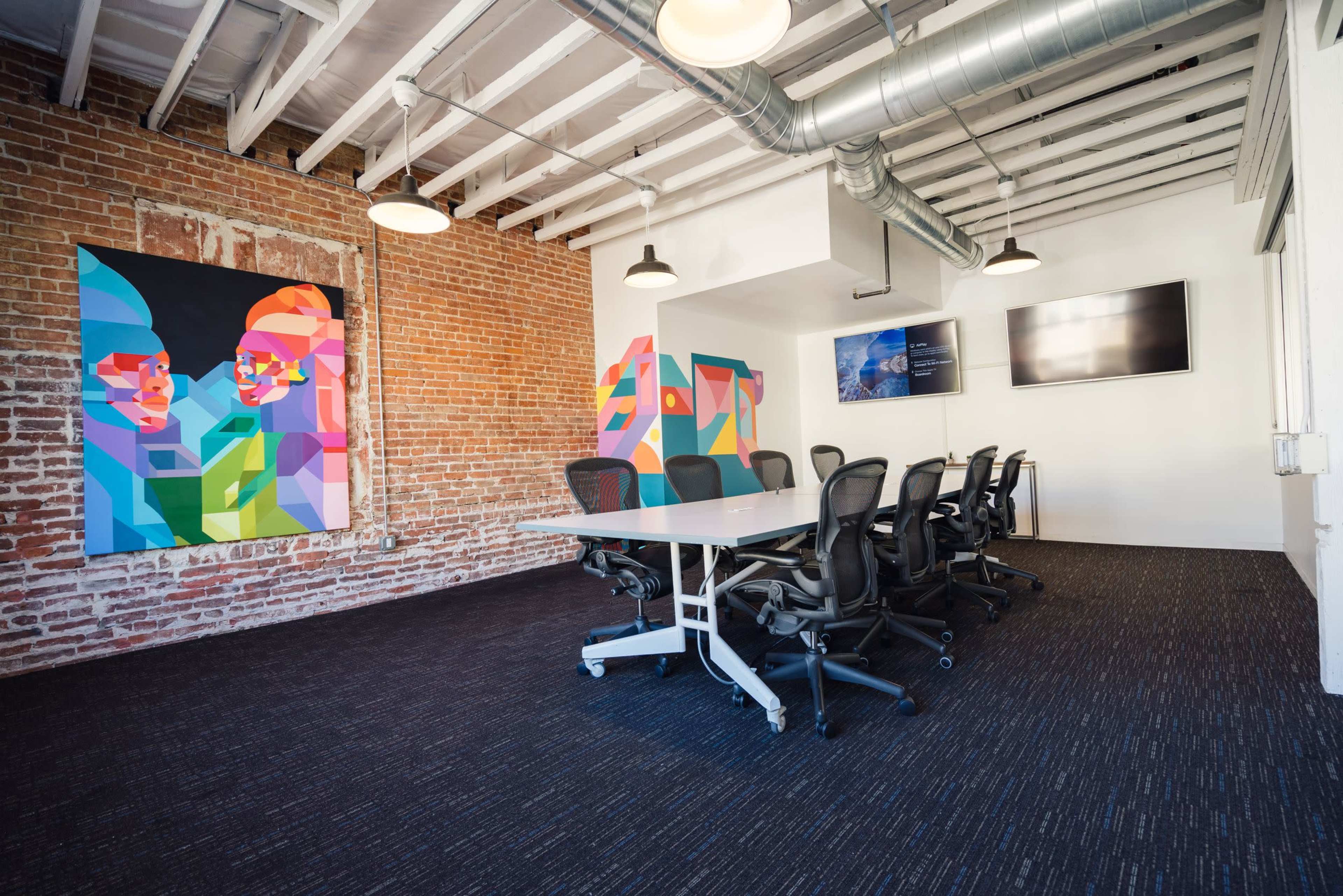 The image shows a modern conference room with a large table, ergonomic chairs, exposed brick walls, and colorful abstract artwork.