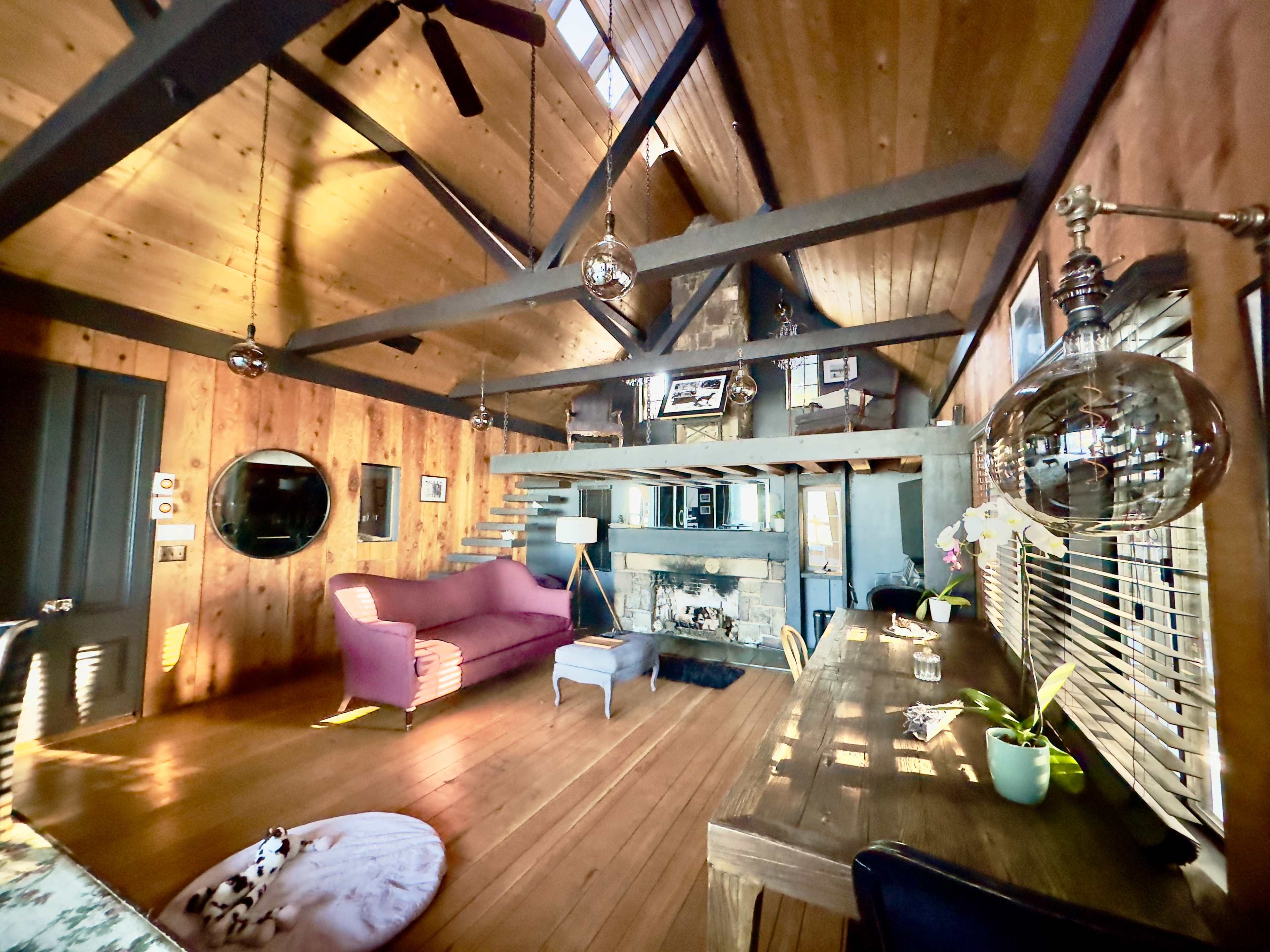 A rustic living space with wooden walls, a vaulted ceiling, a pink sofa, a wooden table, and a cozy fireplace.