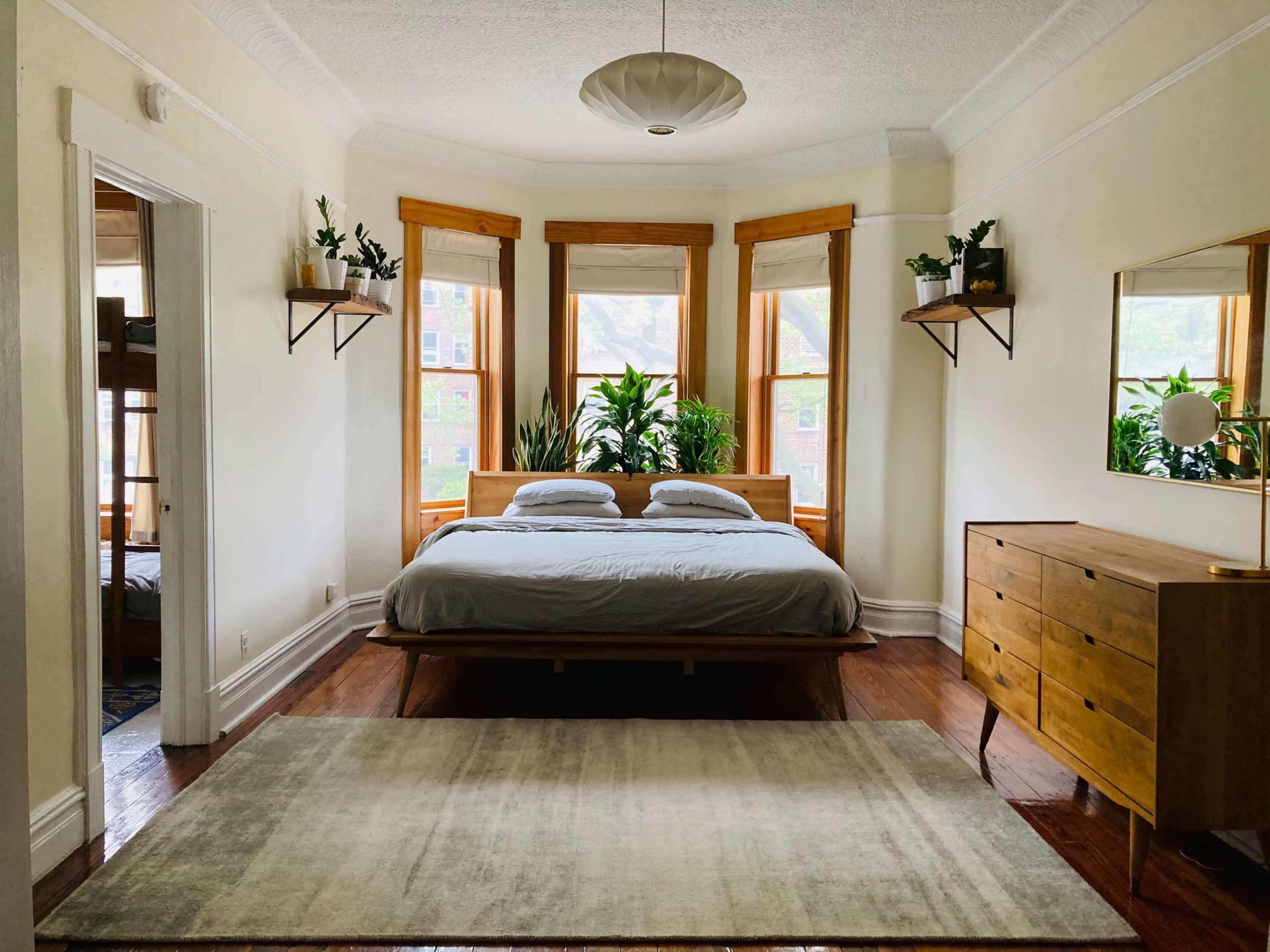 A spacious bedroom features a wooden bed, a dresser, large windows with plants, and light-colored walls.