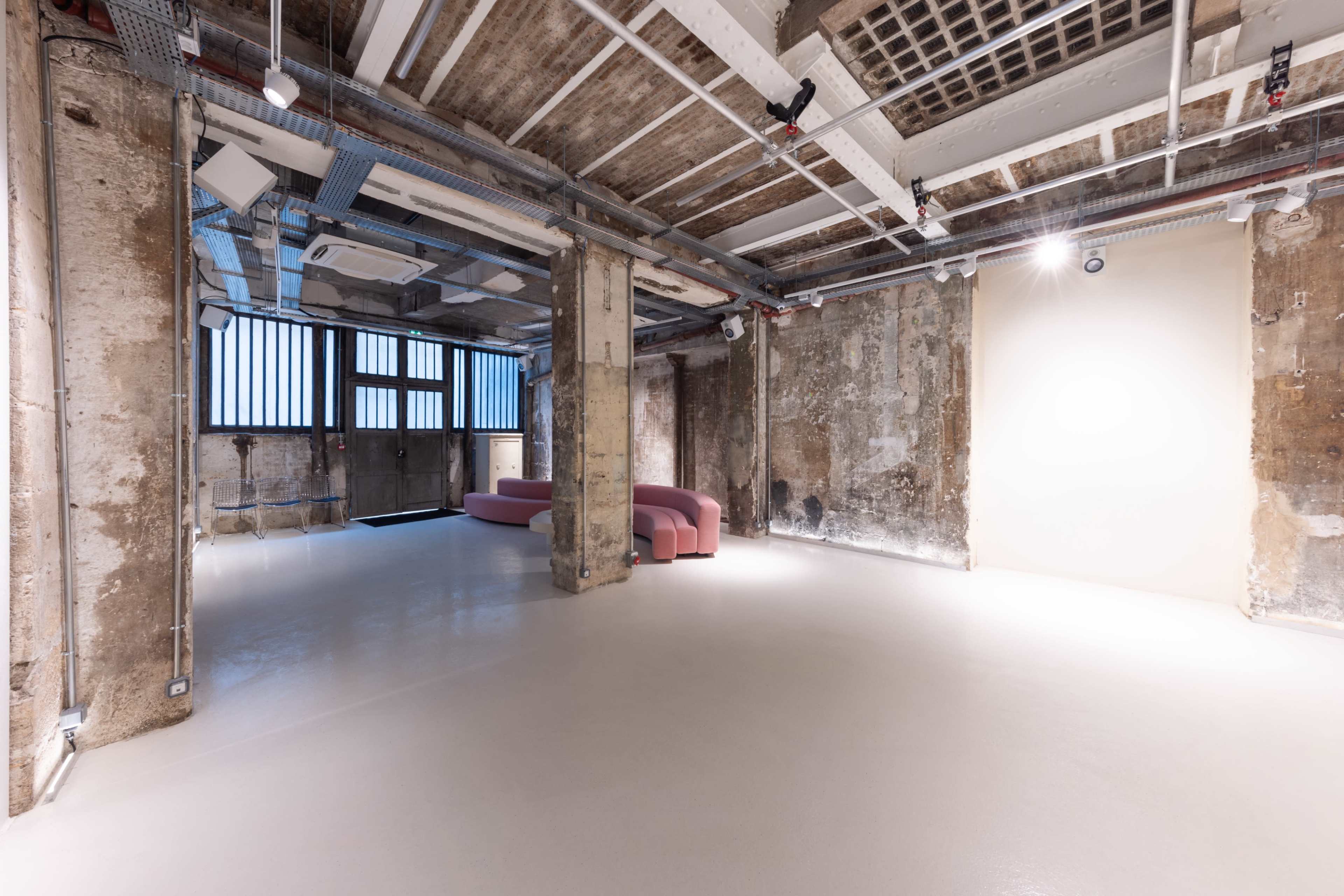 The image shows an industrial-style room with exposed concrete walls, large windows, and a pink curved sofa positioned in the center.