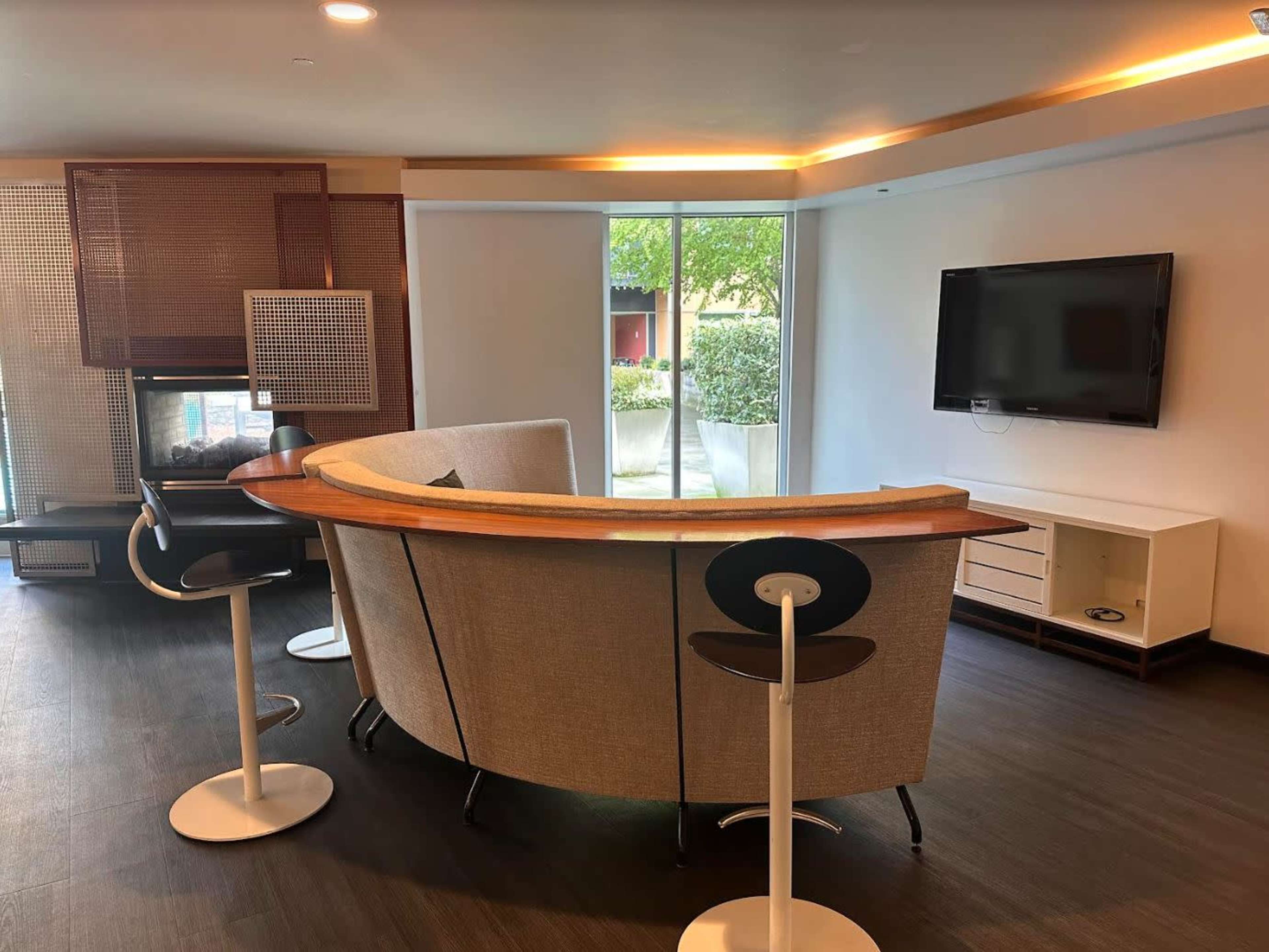 A modern lounge area featuring a curved sofa, bar stools, a television, and large windows with a view of greenery outside.