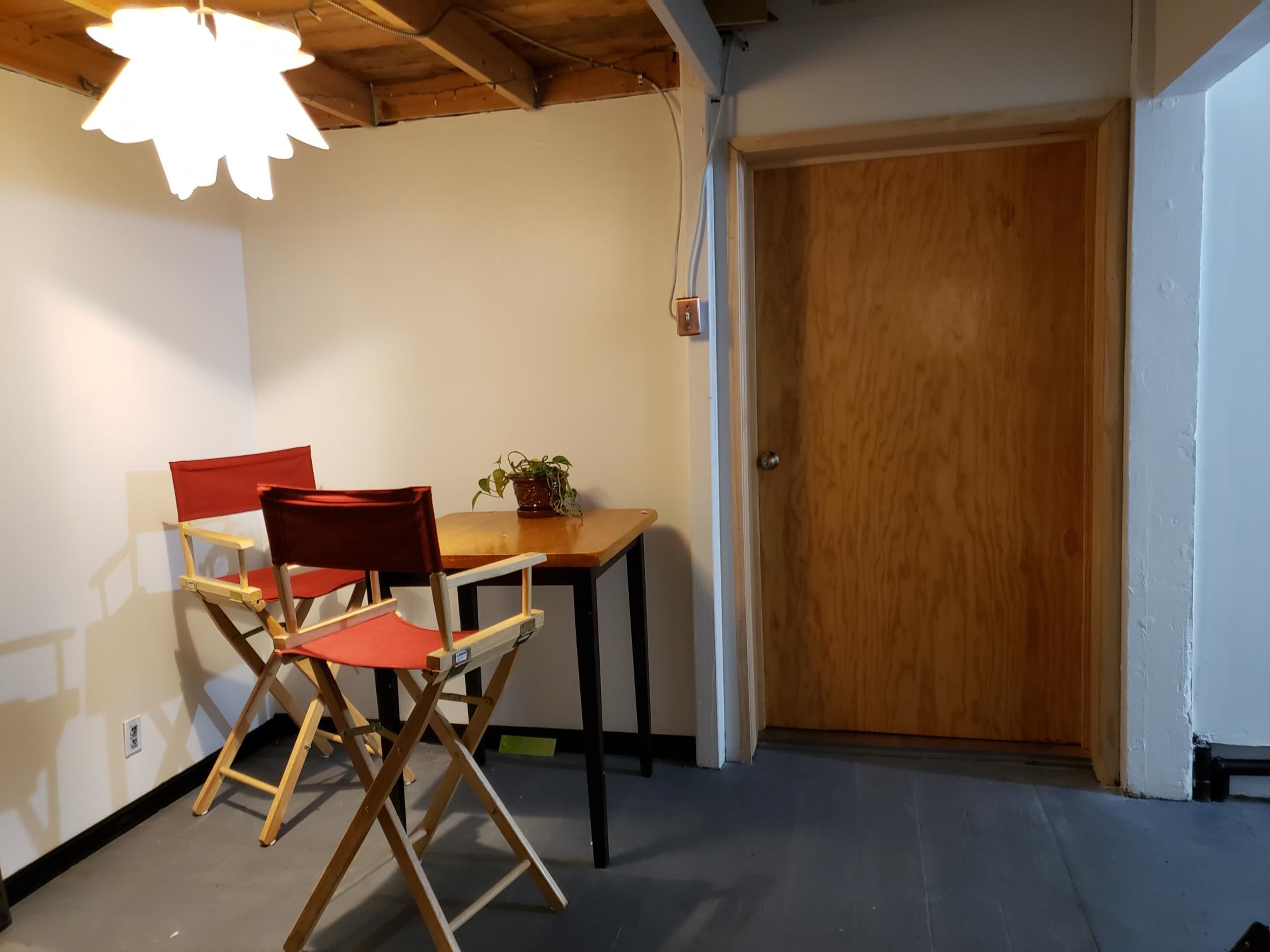 A small room featuring a wooden table with two director-style chairs, a potted plant on the table, and a closed wooden door on one wall.
