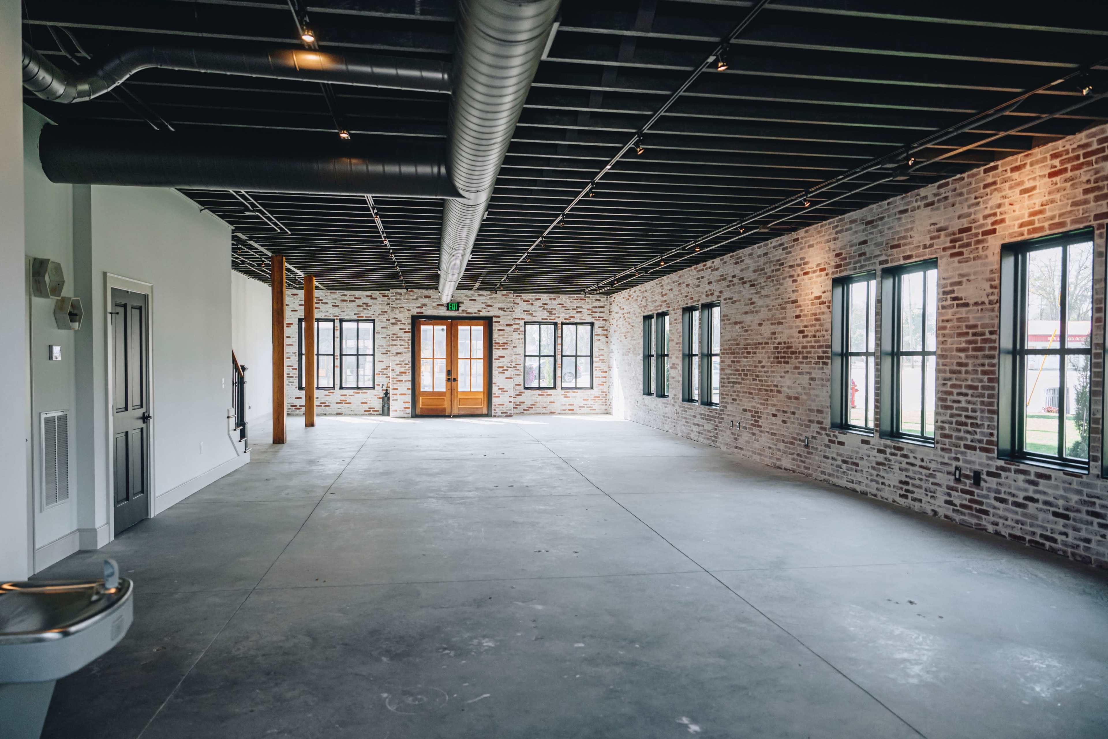 An empty interior space with exposed brick walls, large windows, and a concrete floor.