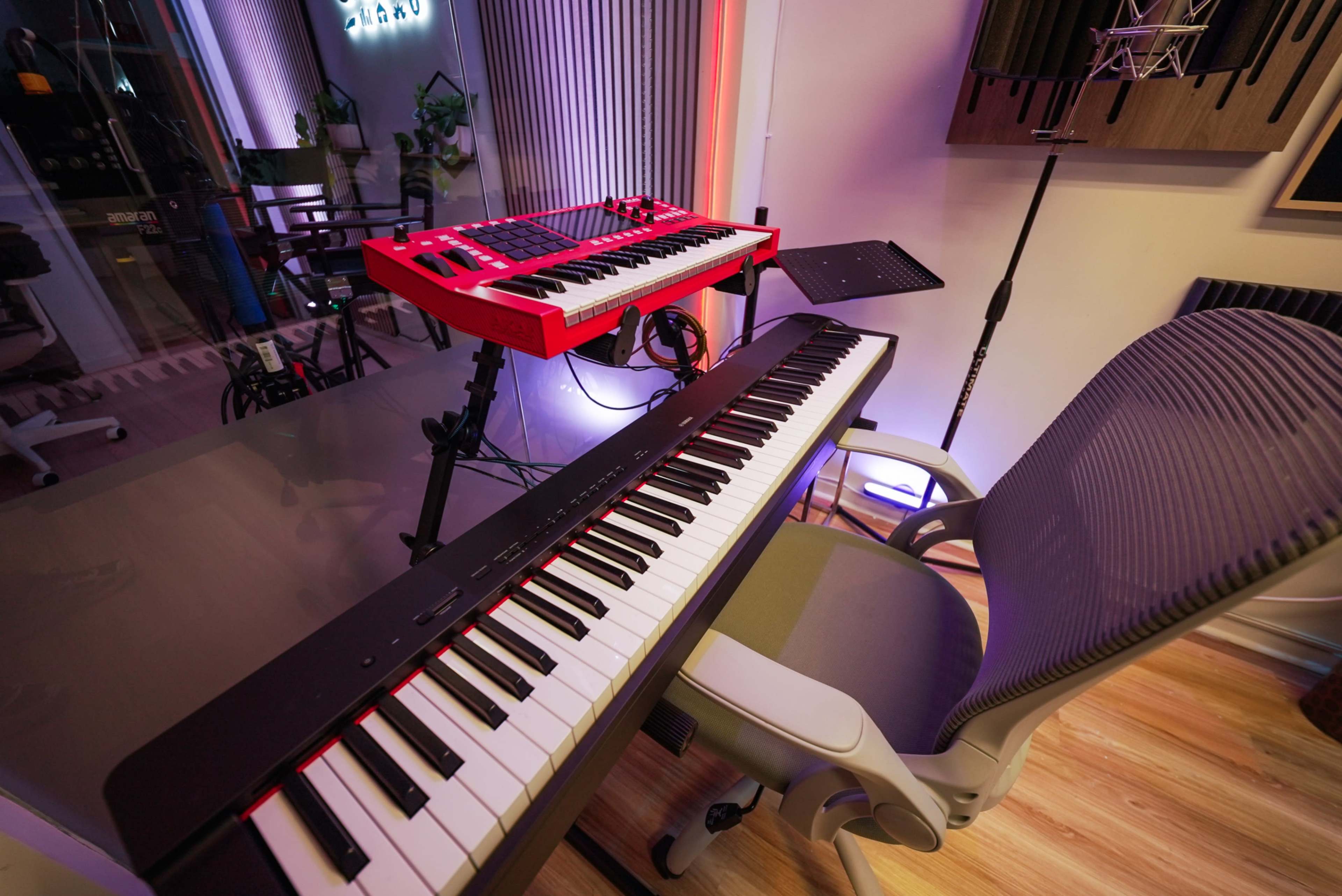 The image shows a music studio setup with two keyboards, a red MIDI controller on a stand, a digital piano on a surface, and an ergonomic chair nearby.