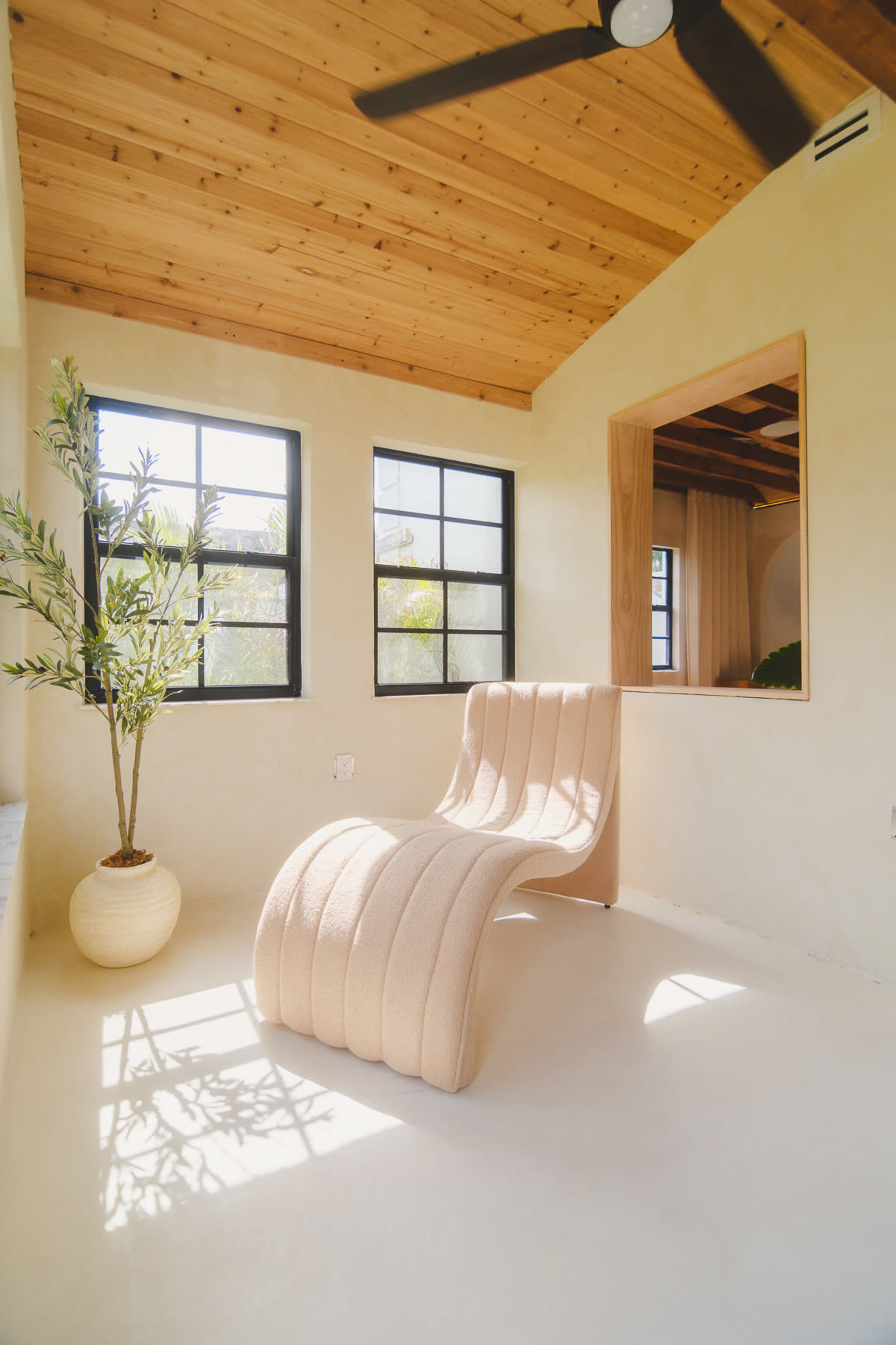 The image shows a minimalist interior with a curved lounge chair beside a large window, accentuated by a potted plant and wooden ceiling.