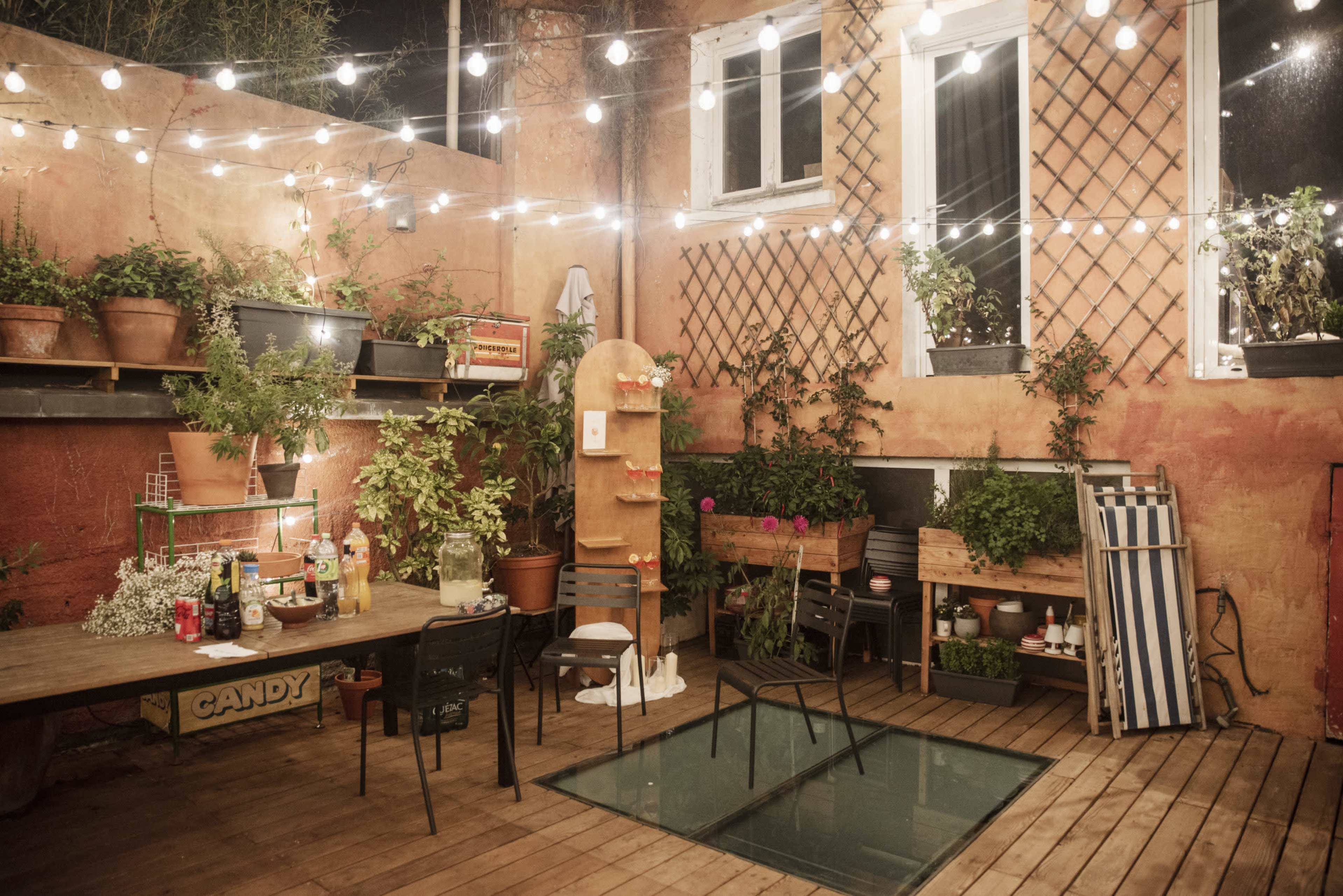 A cozy outdoor patio features a wooden table, chairs, and planters, surrounded by string lights and a glass section in the floor.