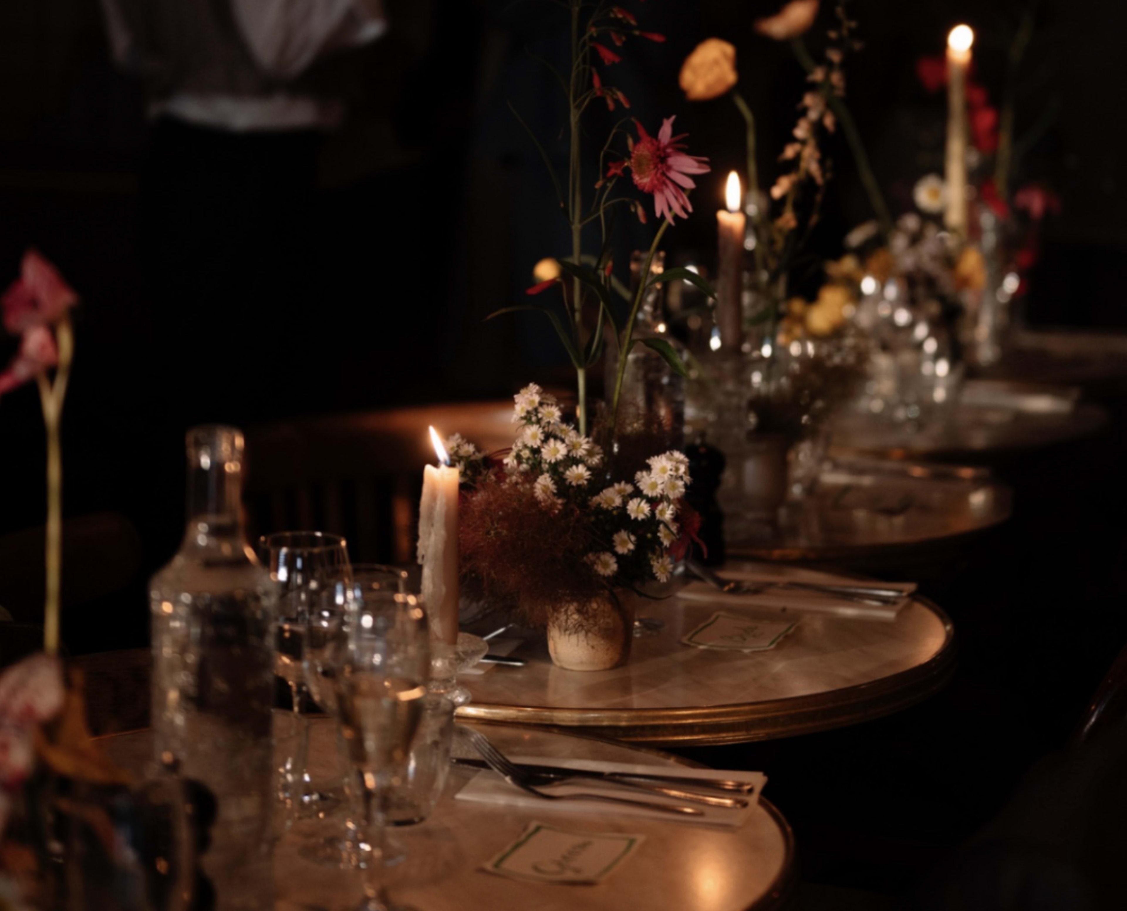 The image shows a dimly lit dining setting with a table adorned with floral centerpieces, candles, and glassware.
