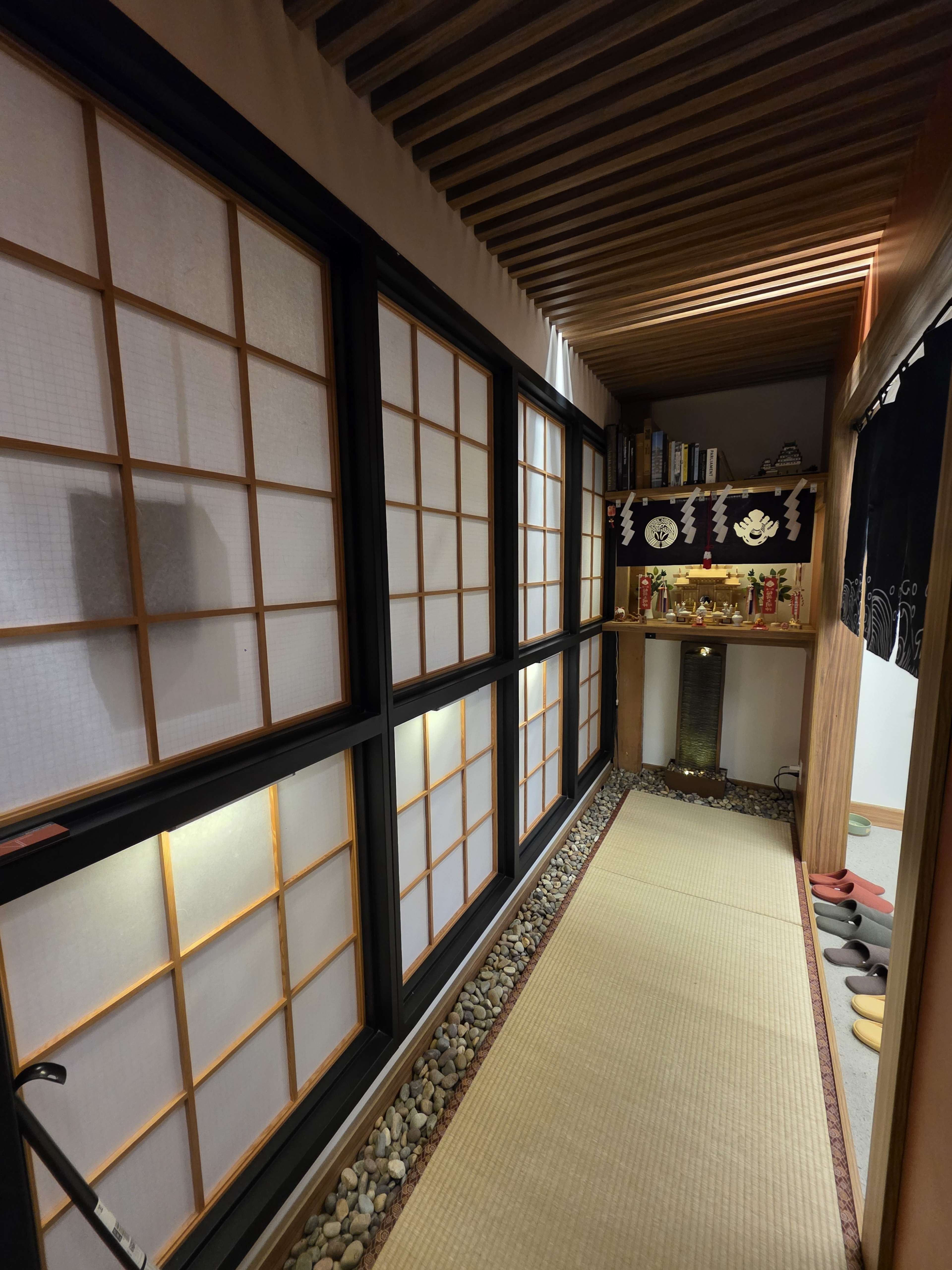 The image shows a narrow hallway with shoji-style windows, wooden beams, and a small altar or display area on one side.