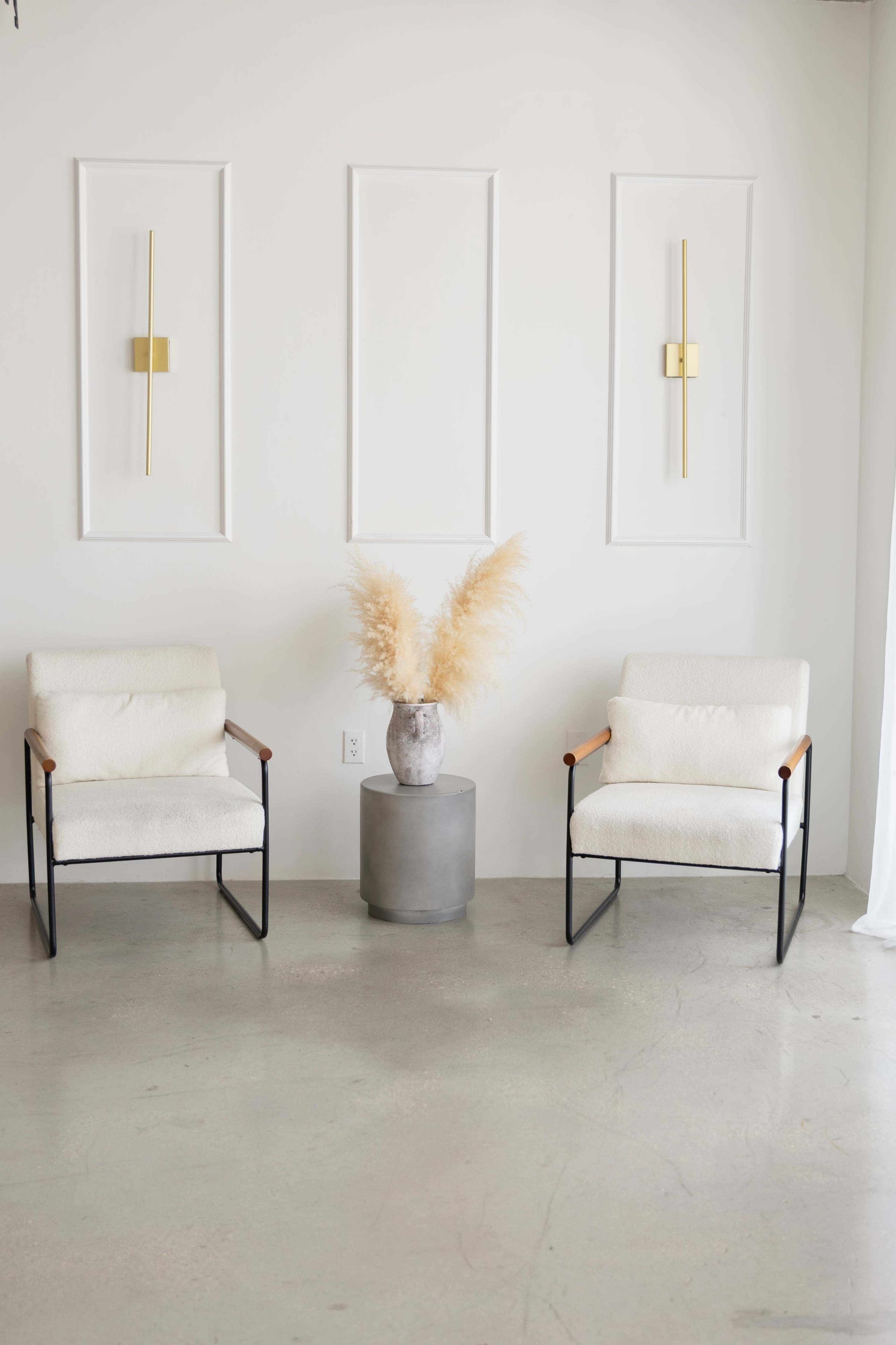 Two modern chairs are positioned opposite each other on a concrete floor, with a minimalistic gray table and a decorative vase containing dried pampas grass placed between them against a plain white wall.