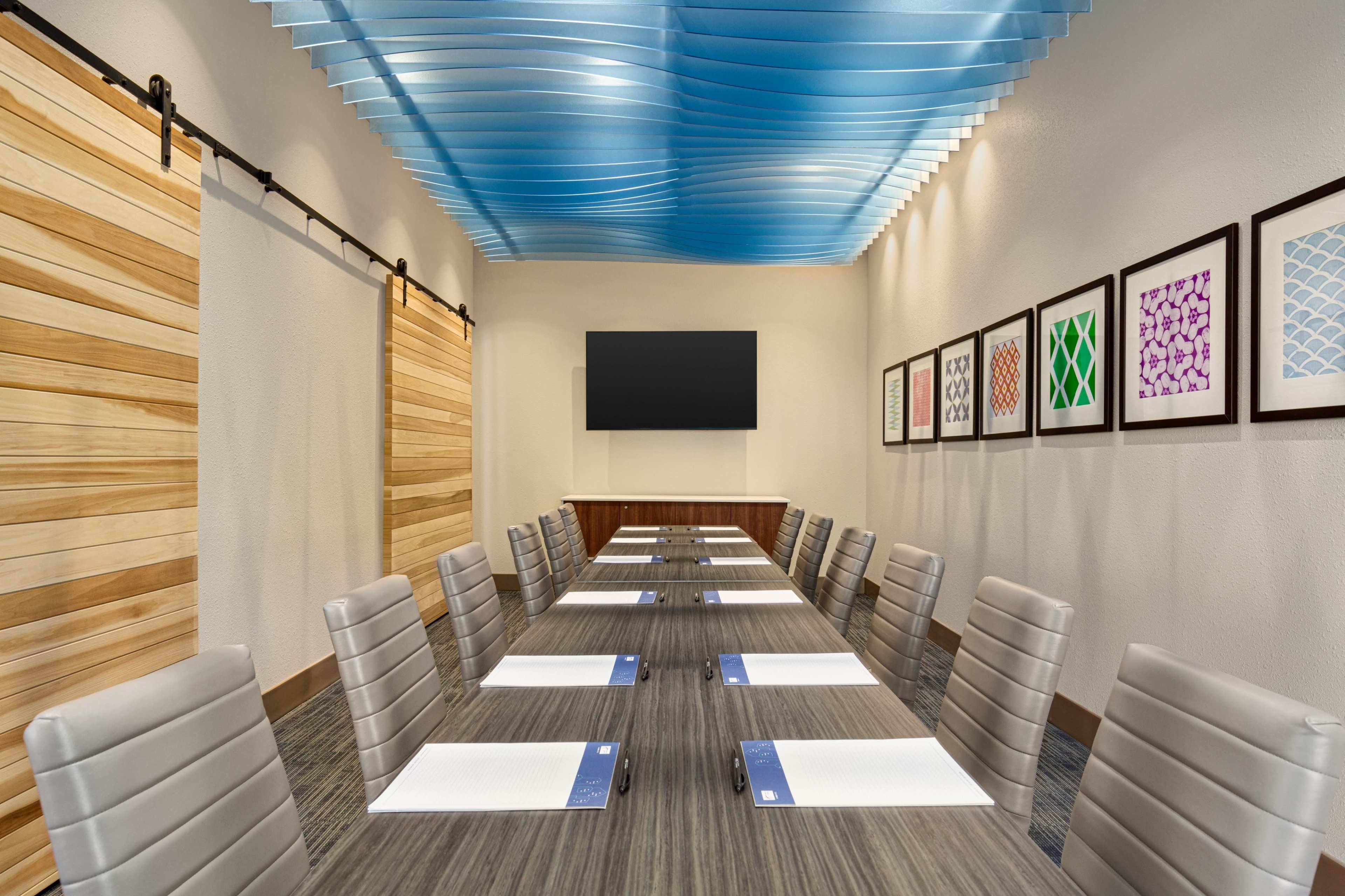 The image shows a modern conference room with a long table, chairs, and wall art, featuring a large screen at one end.