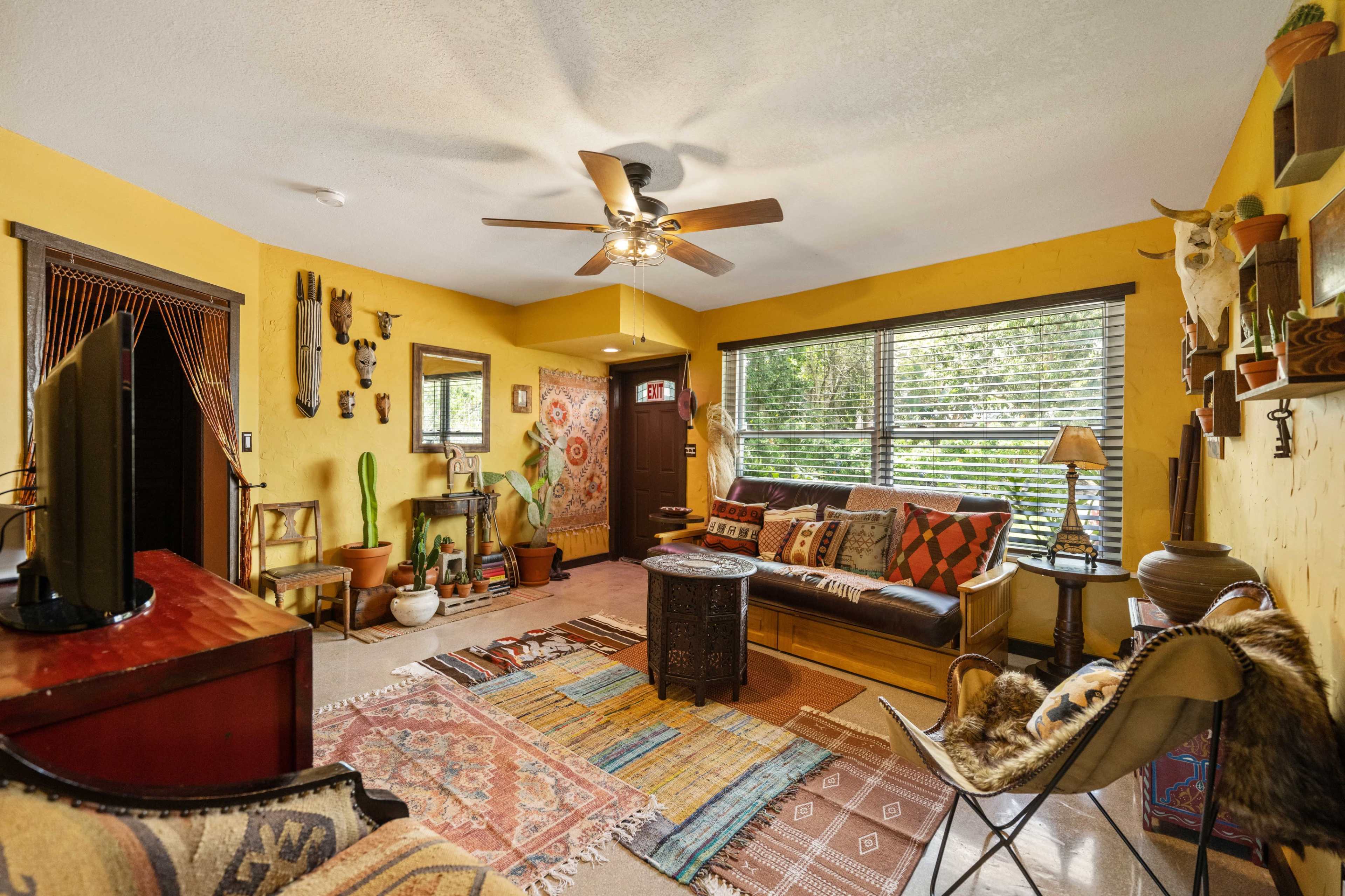 A cozy living room with yellow walls, a ceiling fan, various plants, and colorful textiles arranged on the floor and furniture.