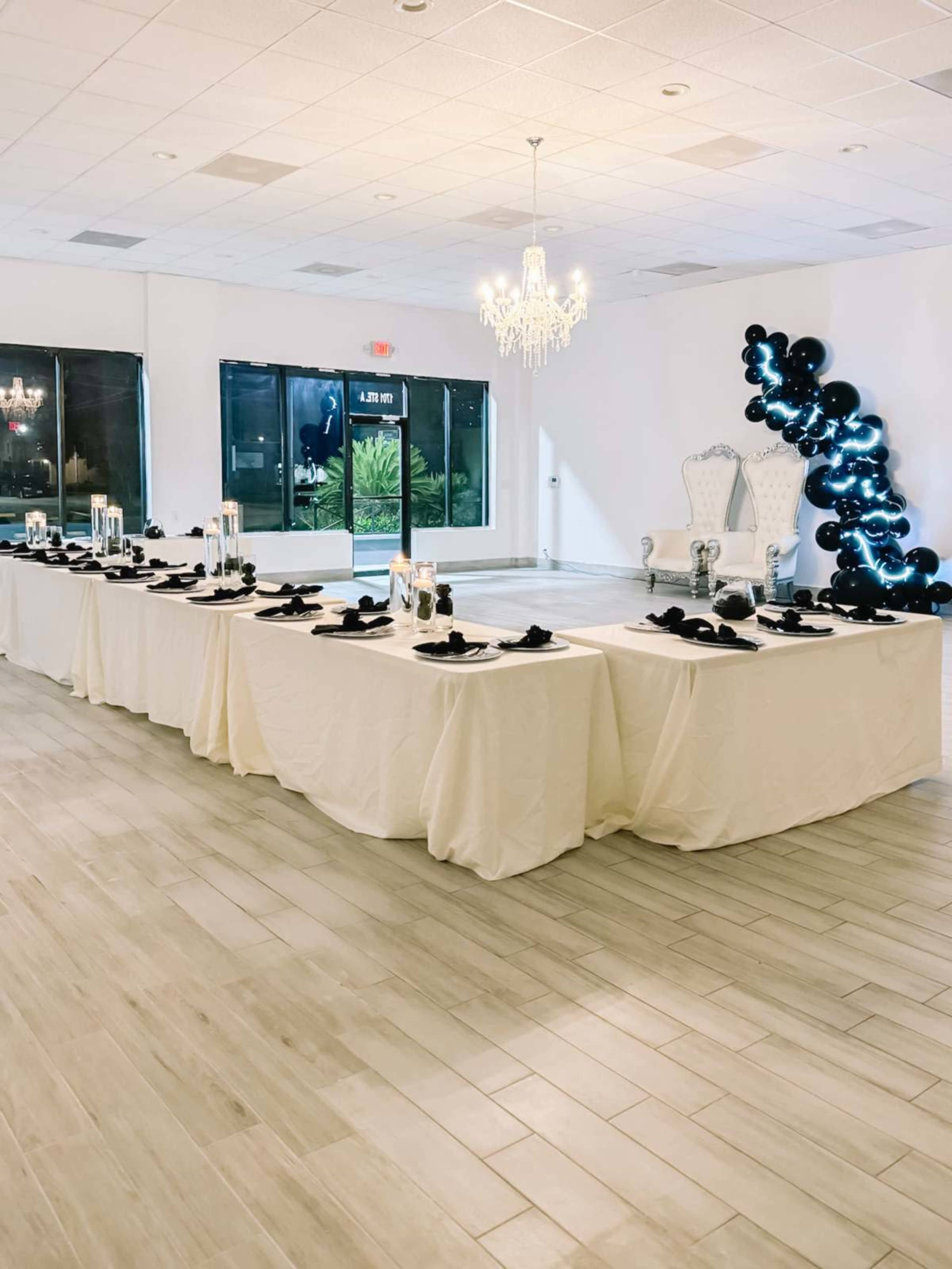 A banquet hall is set up with a long table covered in white tablecloths, black napkins, and decorated with glass candle holders, while a chandelier hangs overhead and a black balloon arch is positioned at one end.