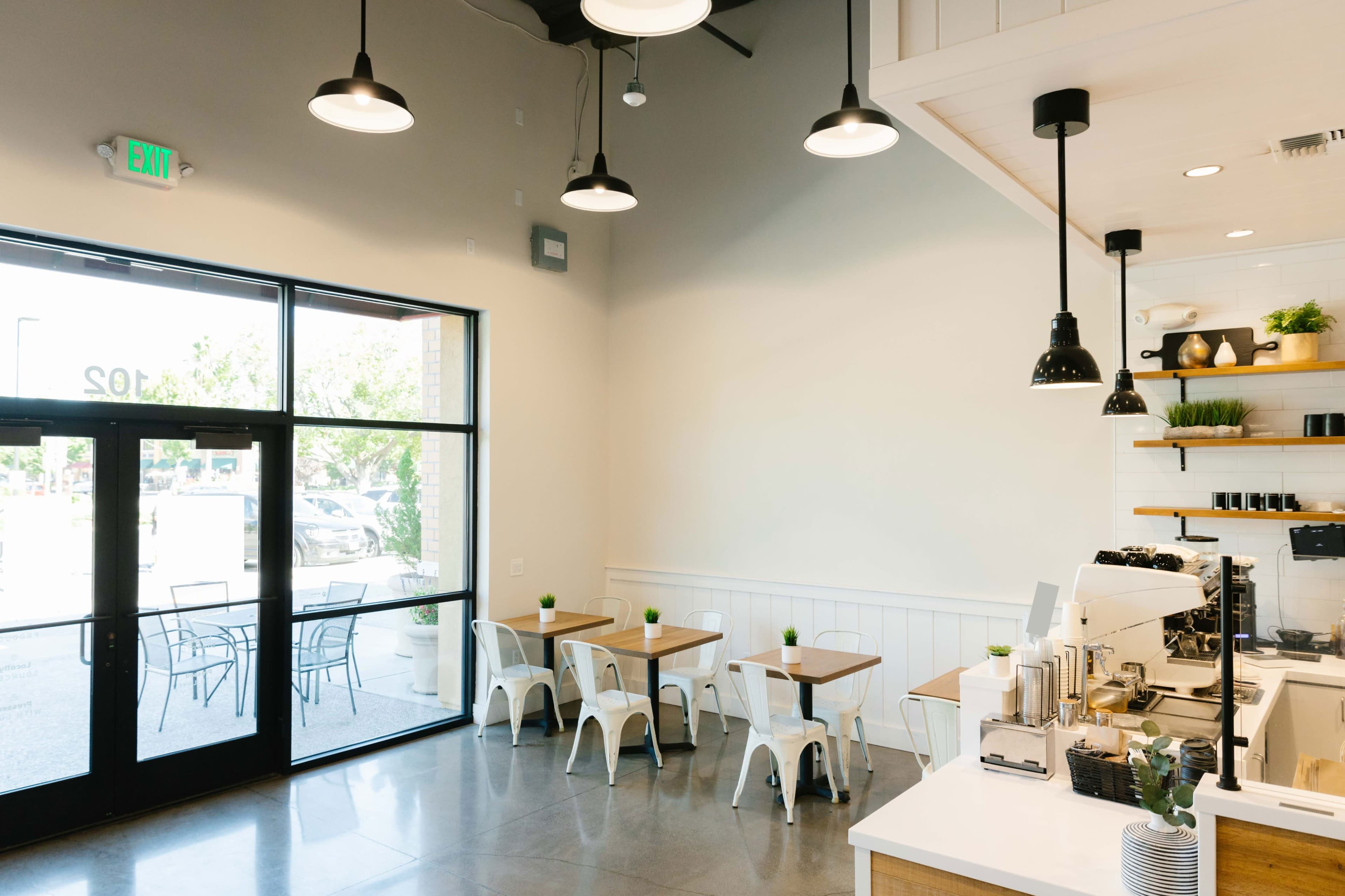 A bright, modern café interior with white walls, metal chairs, and small wooden tables arranged near large windows.