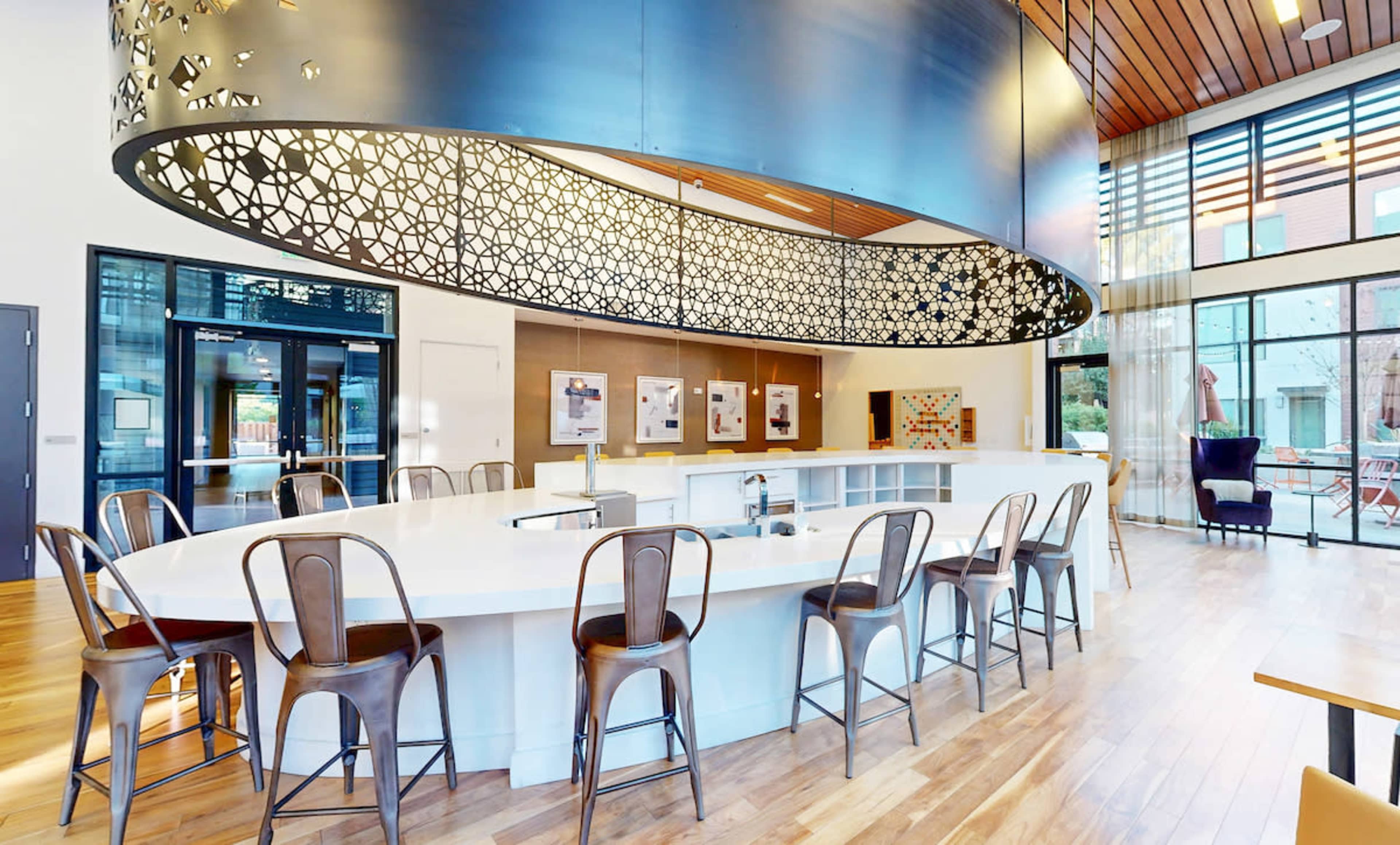 The image shows a modern interior space with a large, curved bar counter surrounded by metal bar stools and decorative overhead features.