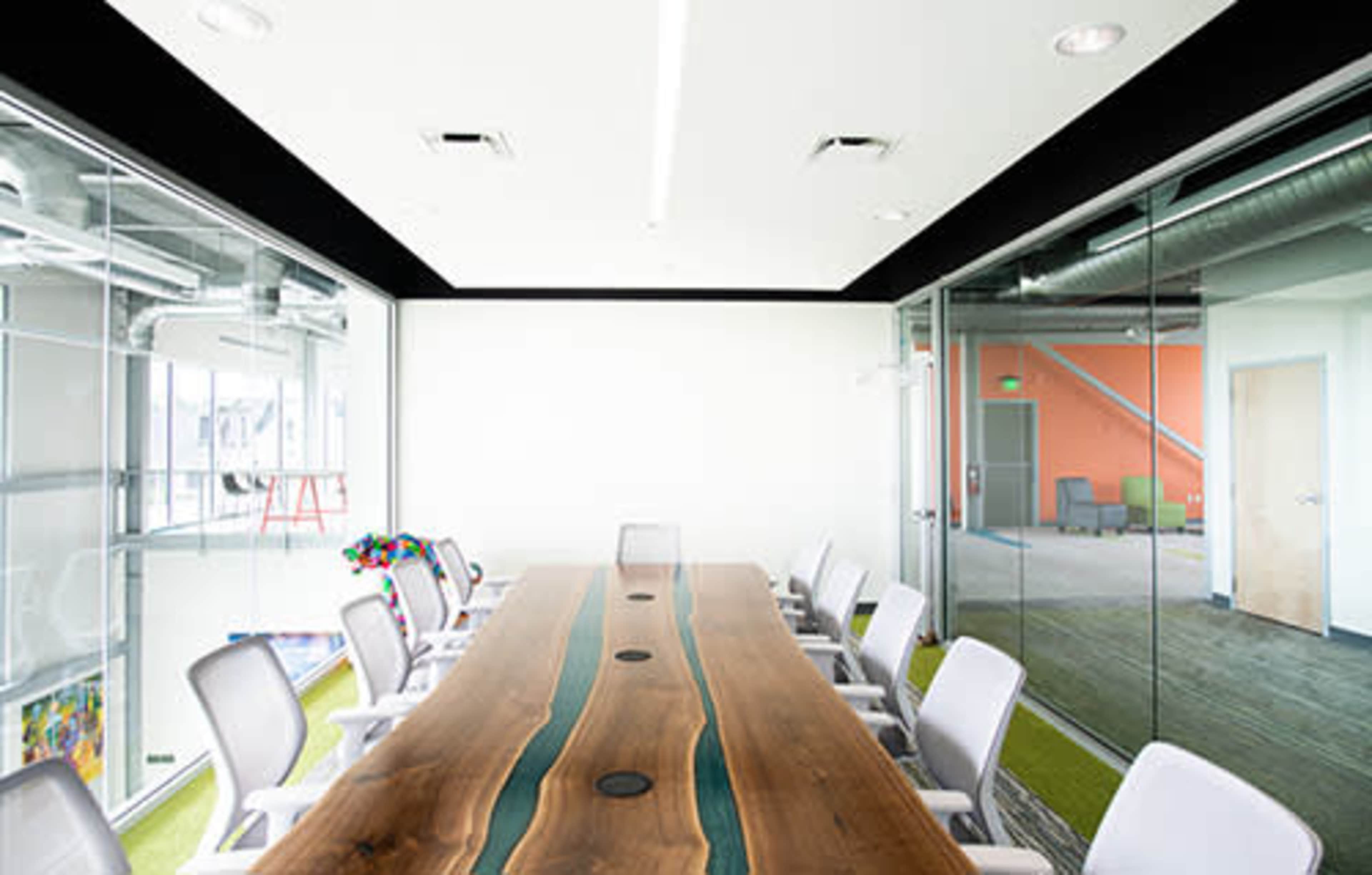 The image shows a modern conference room with a long wooden table featuring a blue epoxy inlay, surrounded by white chairs, next to glass-walled offices.