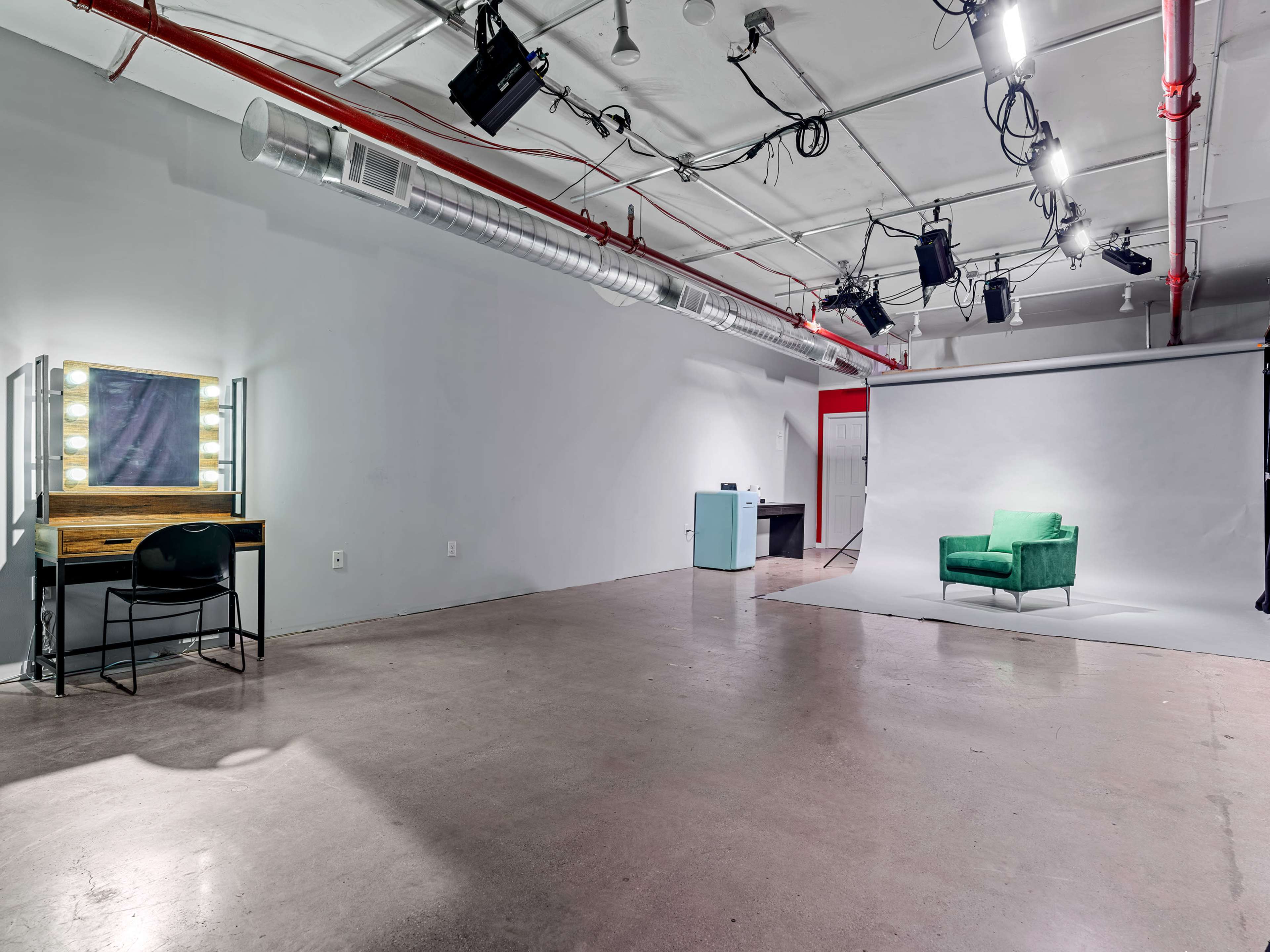 The image shows a spacious studio with gray walls, a green chair on a backdrop, a makeup vanity with lights, and production equipment mounted on the ceiling.