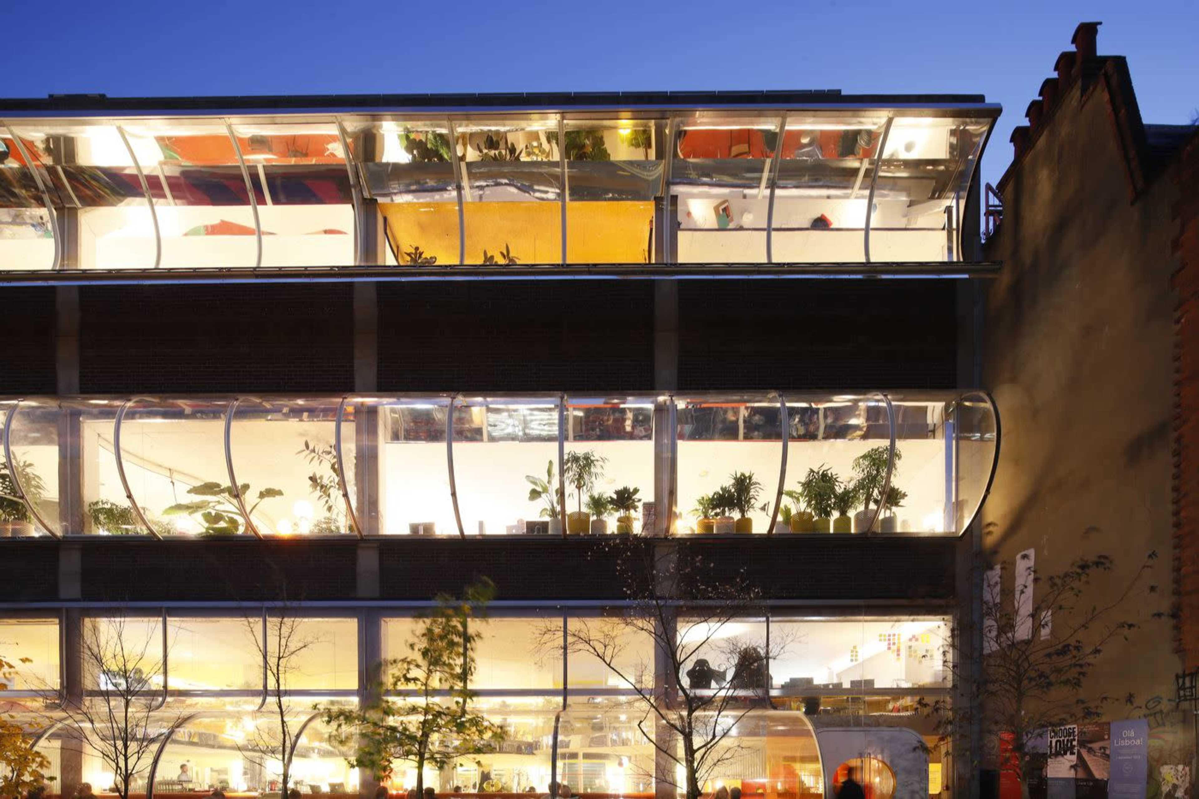 The image shows a multi-story building with large glass windows and curved sections, revealing interior offices filled with plants and warm lighting at dusk.